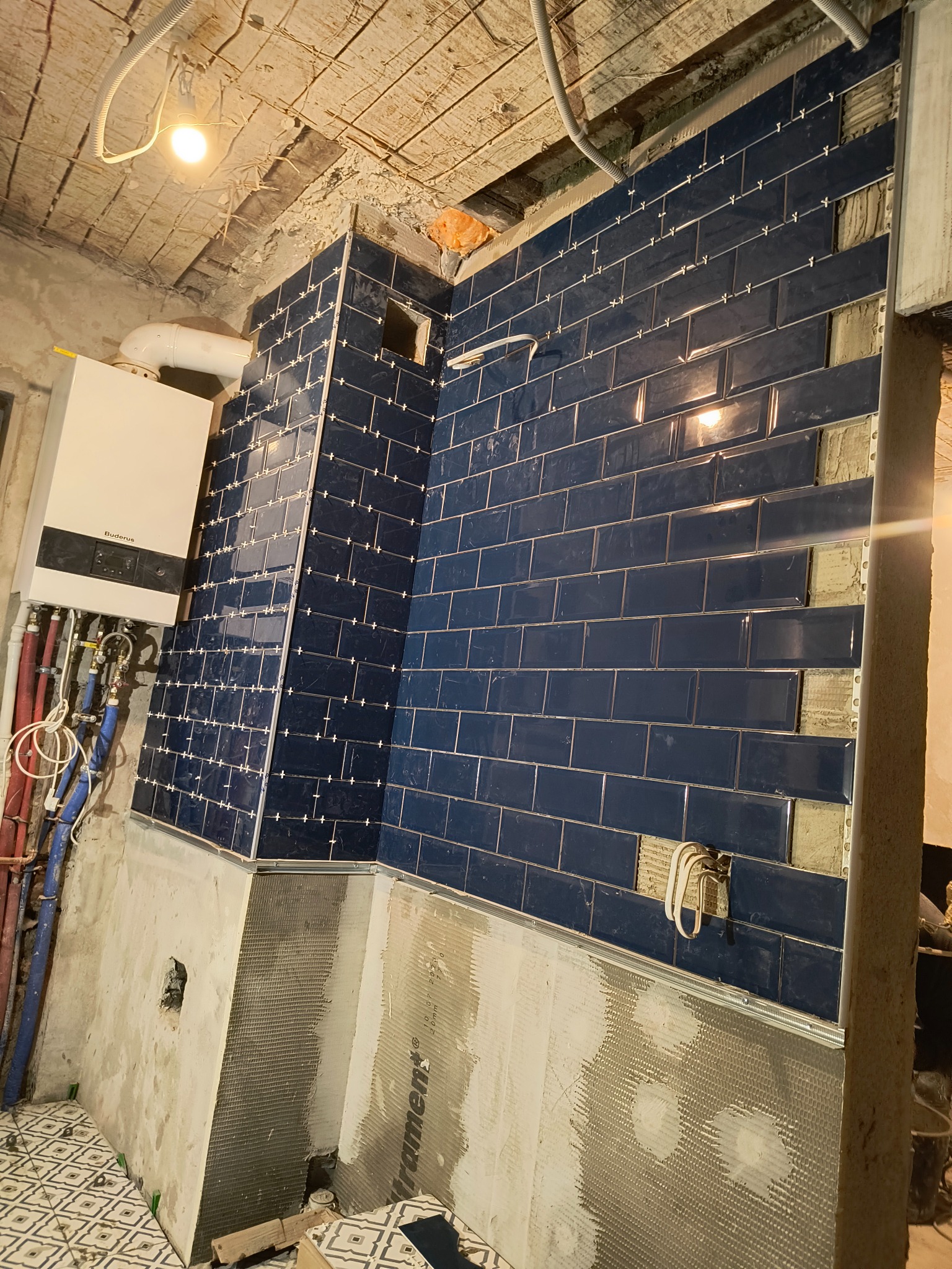 Układanie granatowych płytek typu metro z metalowymi listwami i krzyżykami dystansowymi na ścianie w trakcie prac, widoczny piec gazowy Buderus, surowy sufit i fragment podłogi z mozaiką.