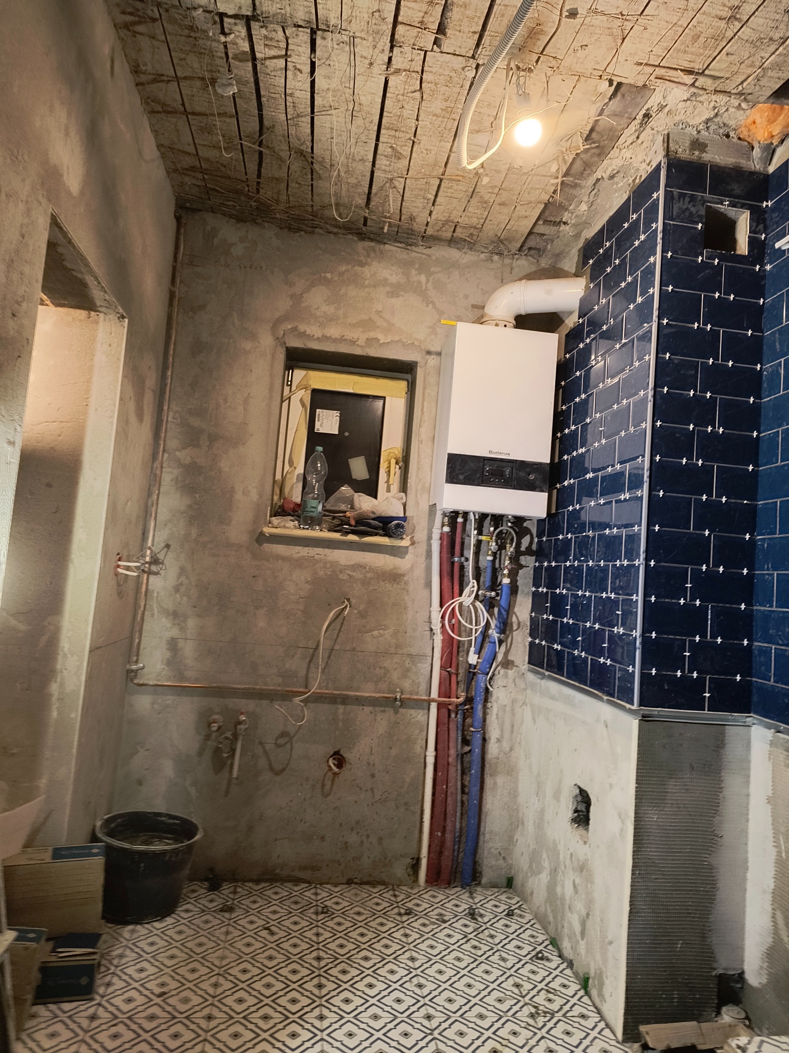 Niedokończona łazienka w trakcie prac: położone ciemnoniebieskie płytki na ścianie, widoczny piec gazowy Buderus z rurami, surowy sufit z odsłoniętymi belkami, podłoga wyłożona wzorzystą terakotą...