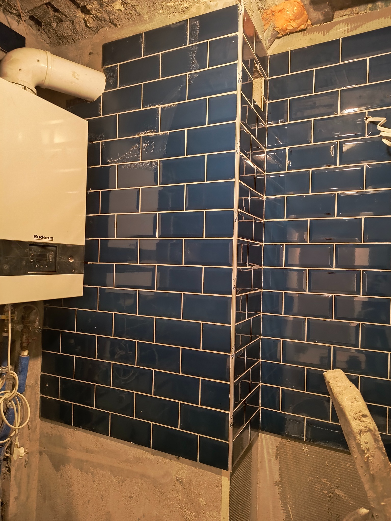 Ściana wyłożona ciemnoniebieskimi, błyszczącymi płytkami w kształcie cegiełek z jasną fugą, widoczny piec gazowy Buderus, instalacja hydrauliczna i fragment surowego sufitu z pianką montażową...