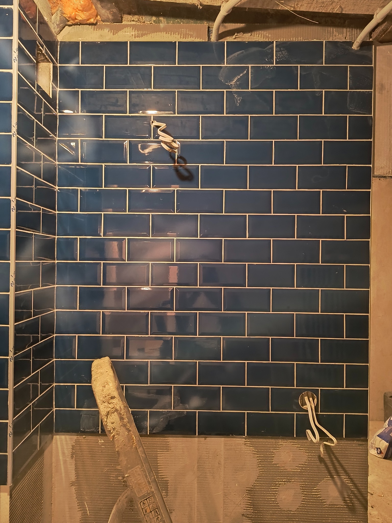 Ściana w trakcie układania błyszczących, granatowych płytek w kształcie cegiełek z jasną fugą, widoczne przewody elektryczne i narzędzie glazurnicze, surowy stan wykończenia.