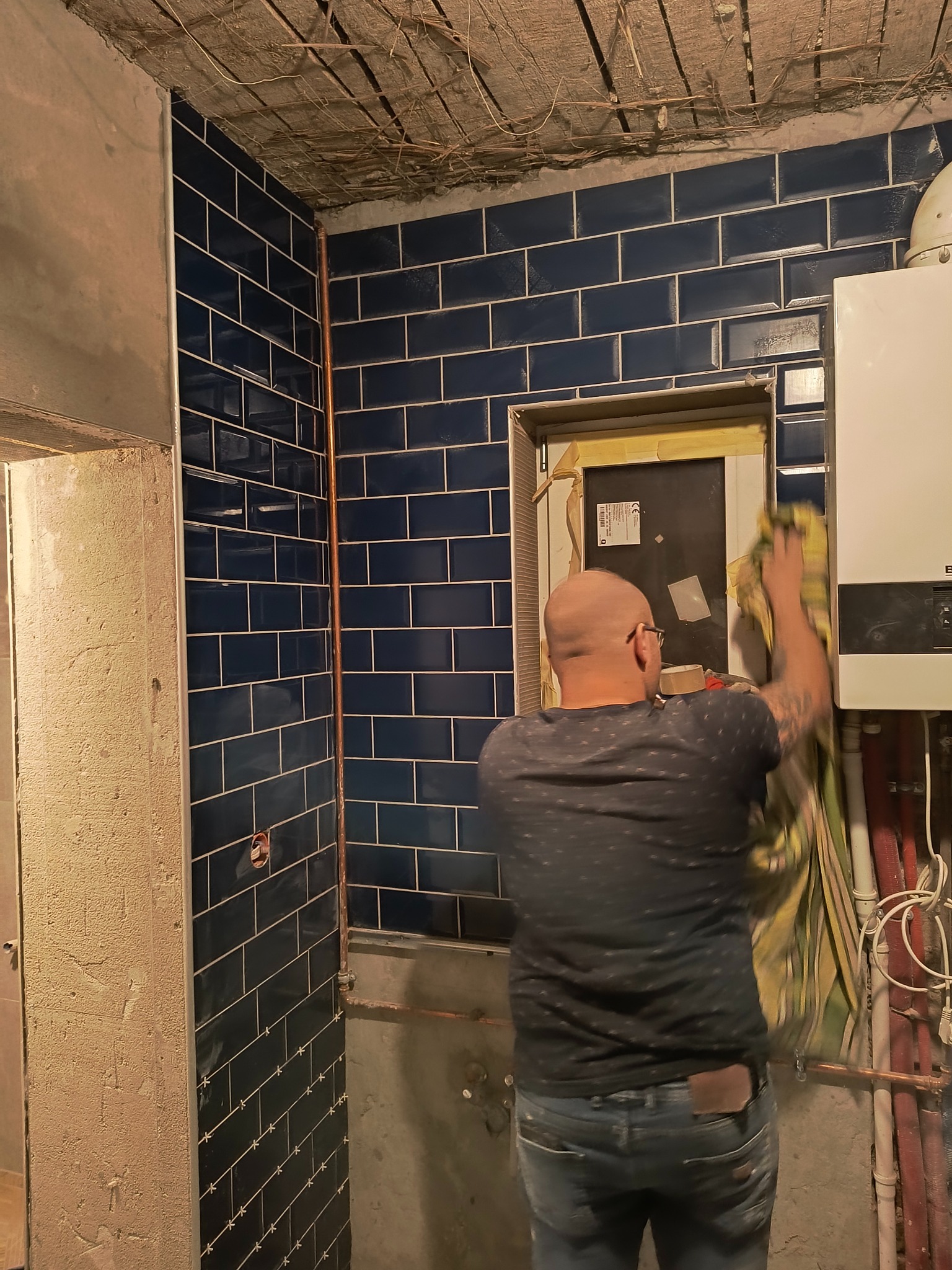 Instalacja granatowych, prostokątnych płytek w łazience, widoczne rury miedziane i fragment bojlera, mężczyzna czyści płytki szmatką, surowy sufit przed wykończeniem.