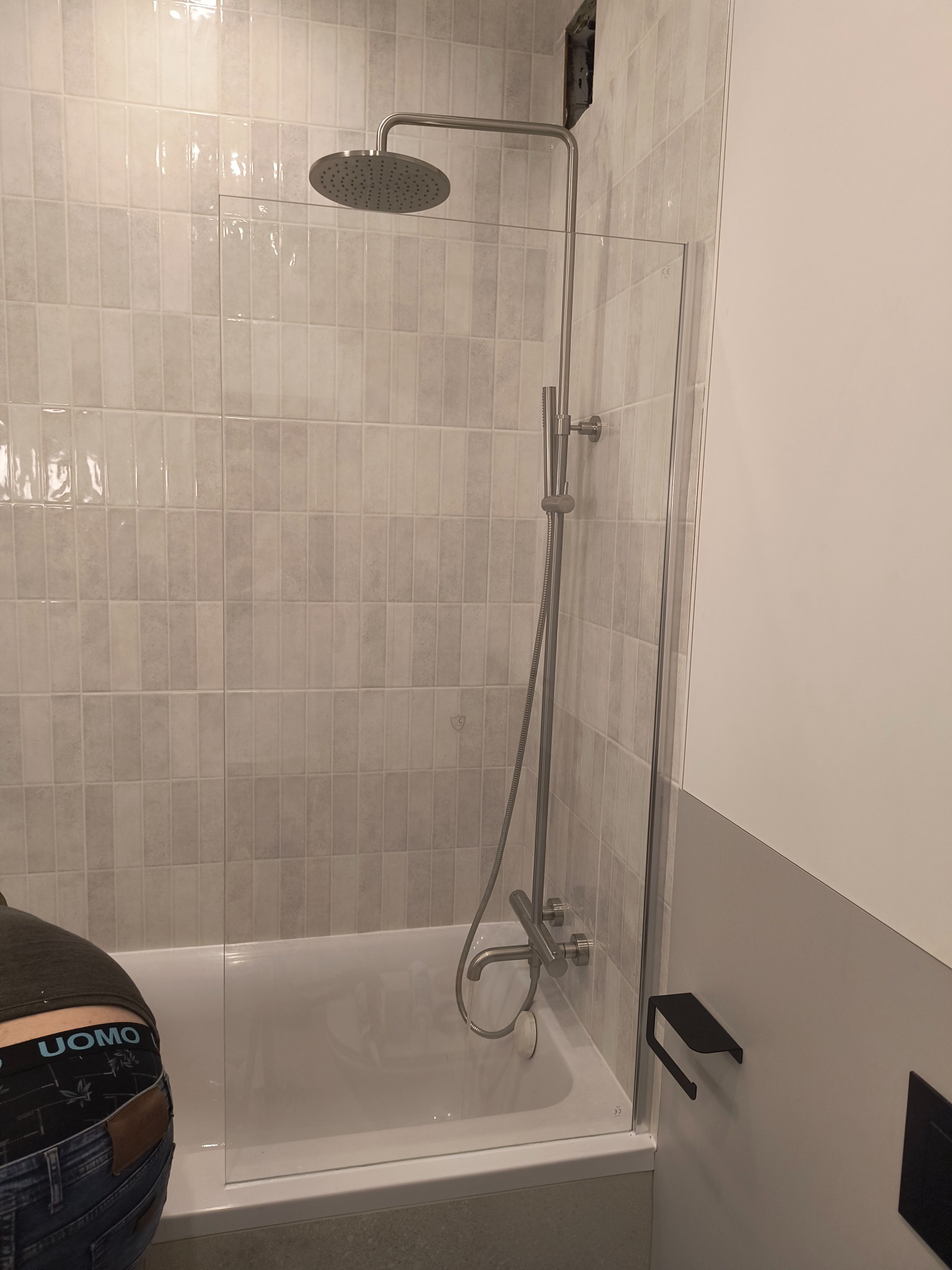 Szklany parawan nawannowy z chromowaną armaturą prysznicową na tle ściany z płytek w odcieniach szarości; widoczny fragment sylwetki osoby w jeansach i bieliźnie z napisem UOMO.