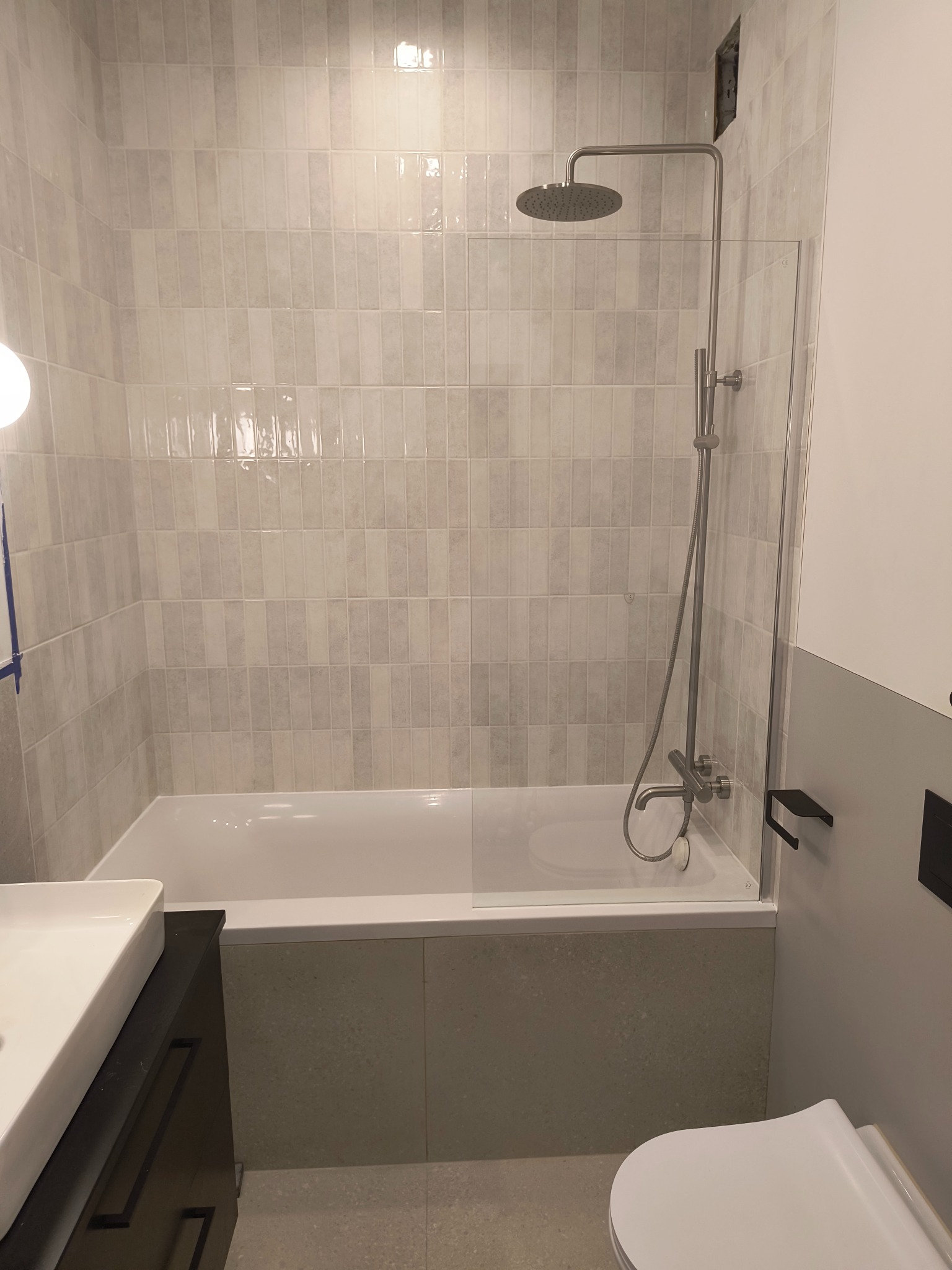 Wykończona łazienka z wanną obłożoną płytkami imitującymi kamień, szklanym parawanem, armaturą w kolorze chromu oraz umywalką z szafką w kolorze ciemnego brązu.