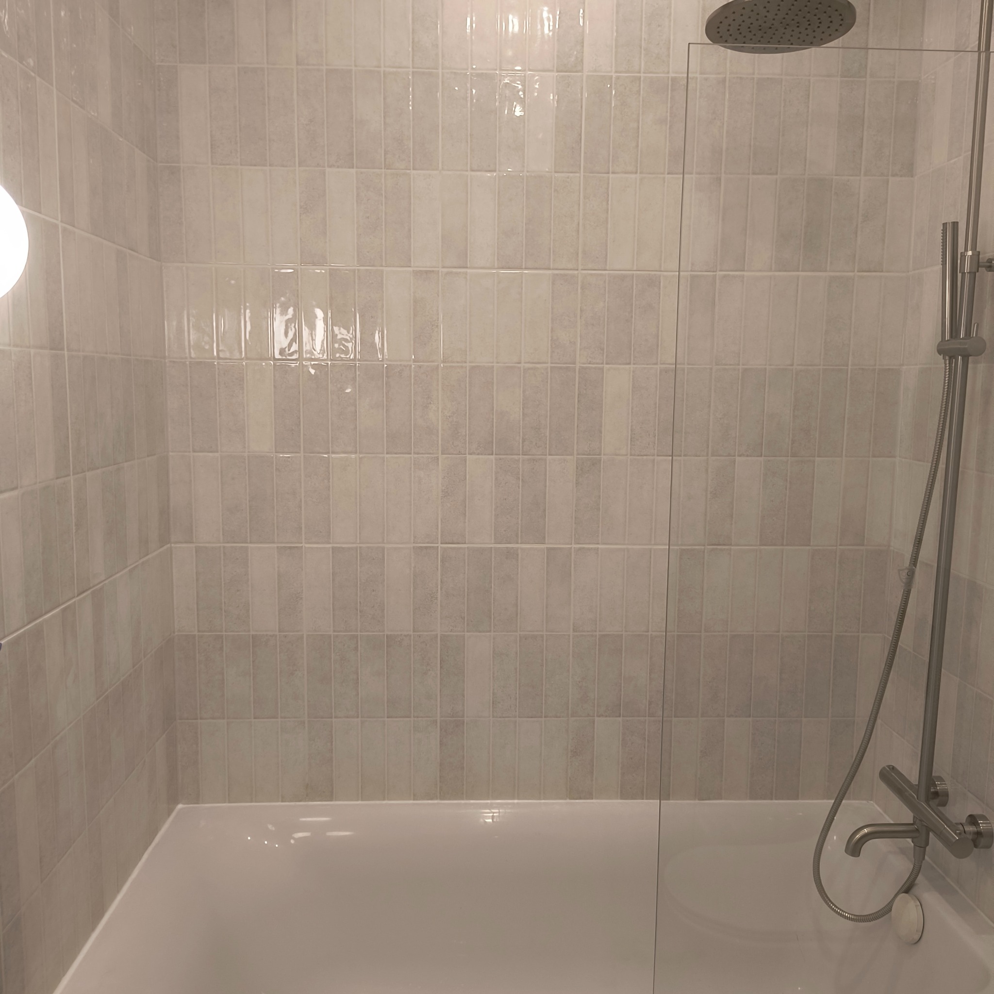 Wykończona łazienka z wanną, szklanym parawanem i baterią prysznicową z deszczownicą, wyłożona jasnymi, pionowymi płytkami imitującymi cegiełki.