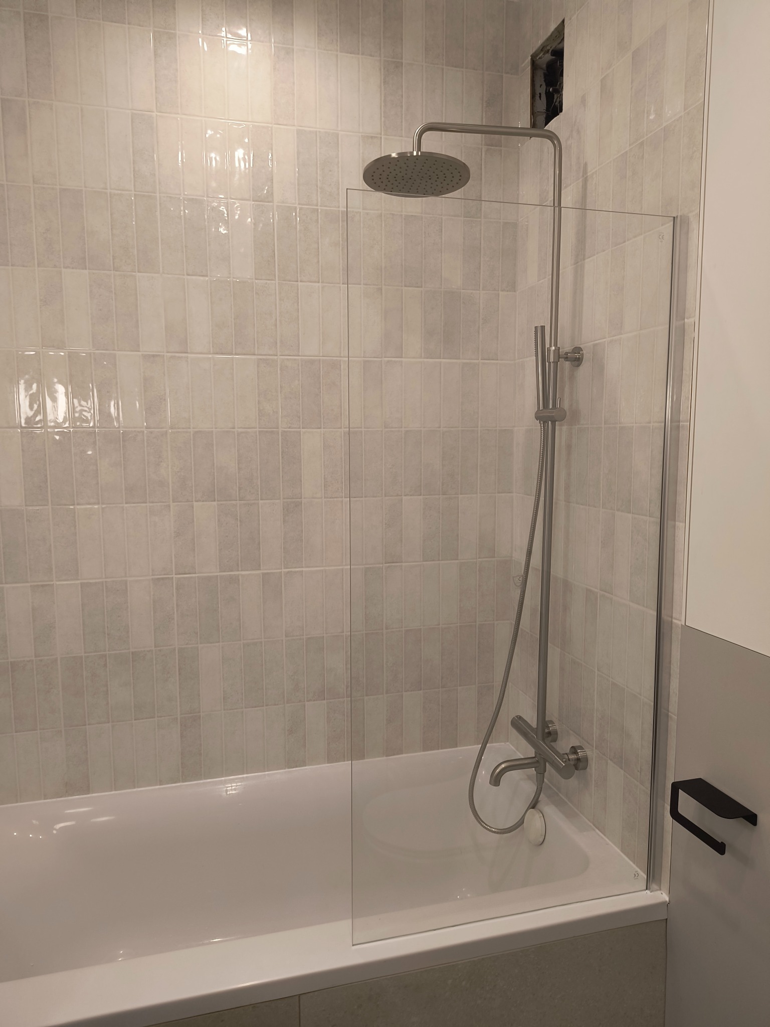 Wykończona łazienka z wanną, szklanym parawanem nawannowym i deszczownicą; widoczna faktura płytek w odcieniach szarości i beżu, brakująca część zabudowy nad prysznicem.