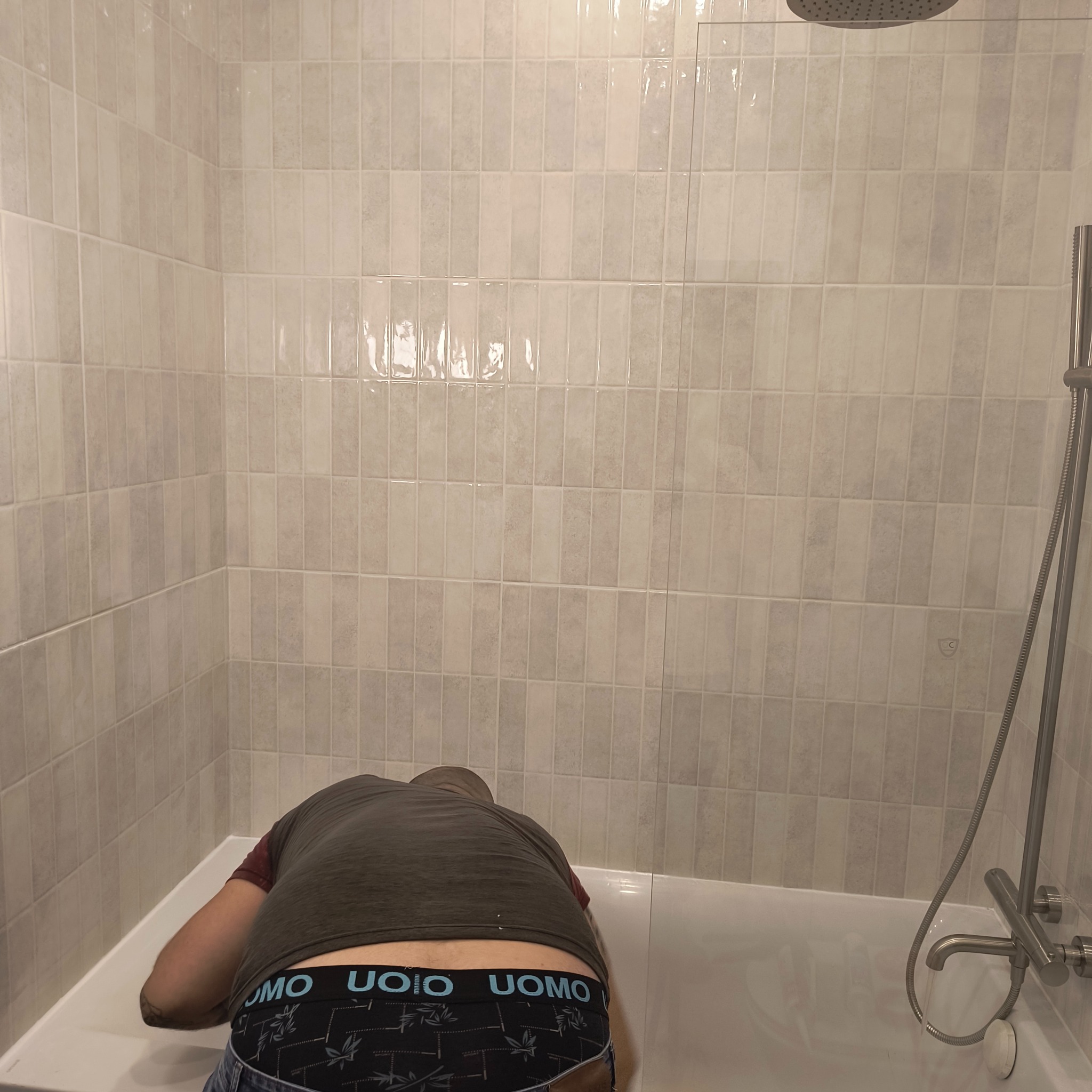 Osoba pochylona nad wanną w trakcie prac wykończeniowych, widoczne płytki w odcieniach szarości i beżu na ścianach, bateria prysznicowa.