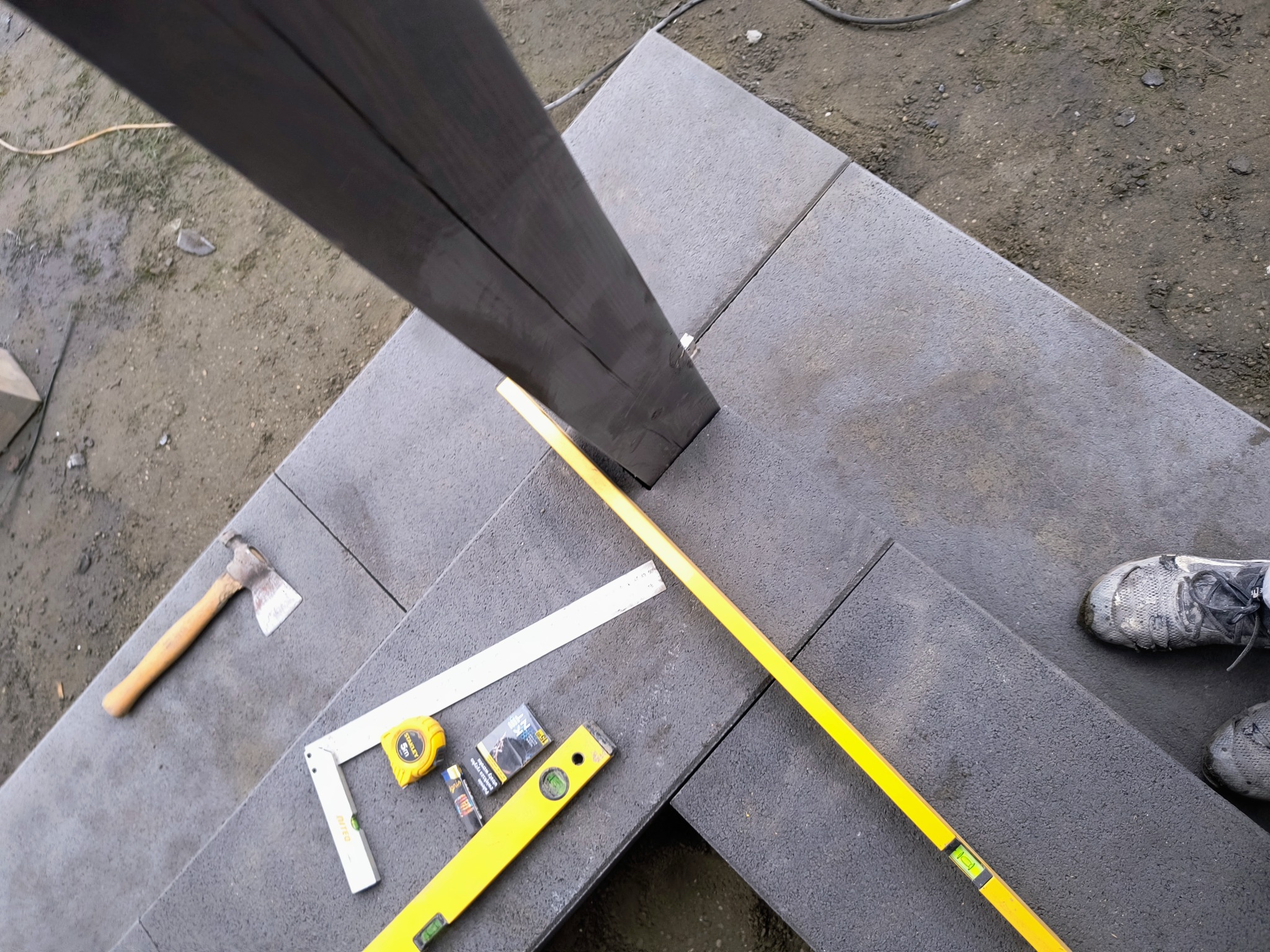 Montaż szarej kostki brukowej z użyciem poziomicy, kątownika, miarki zwijanej, siekiery i drewnianego słupa nośnego; widoczne robocze buty