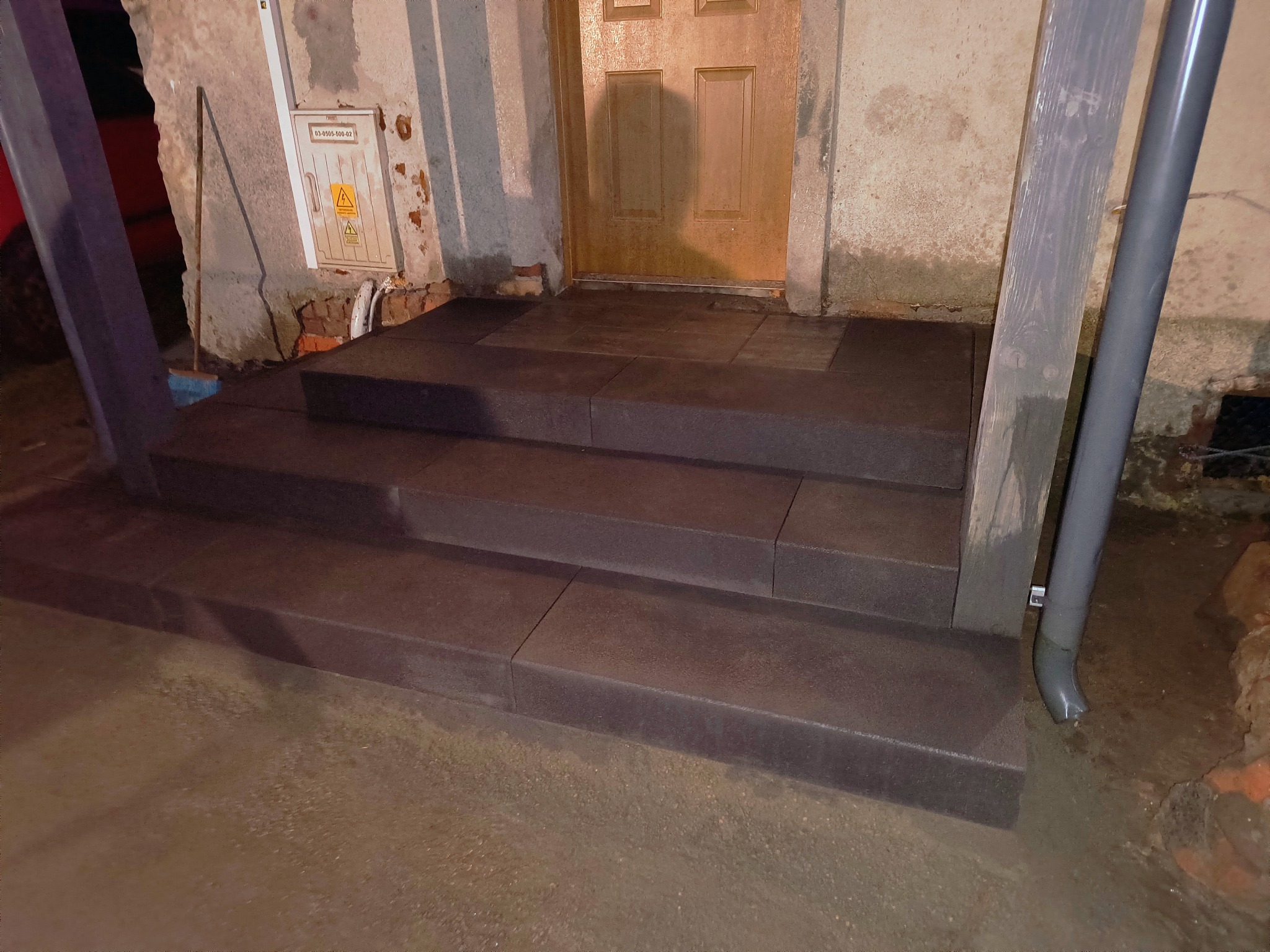 Nowe, ciemnoszare, betonowe schody wejściowe prowadzące do drzwi, z widocznym fragmentem elewacji i rury spustowej. Świeżo położone płyty chodnikowe na podłożu.