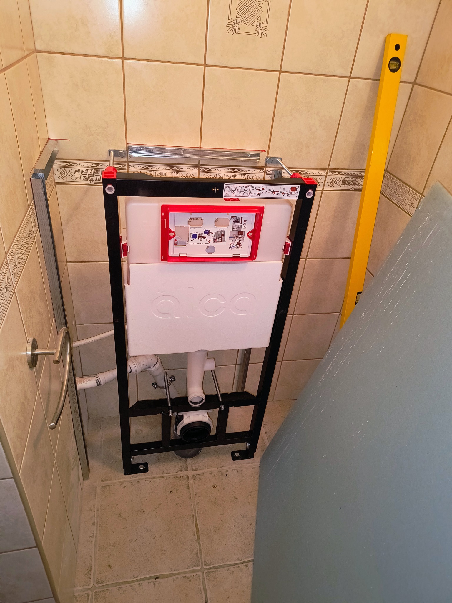 Stelaż podtynkowy WC marki Alca, zamontowany w łazience z beżowymi płytkami, widoczny poziomica i fragment płyty karton-gips, etap instalacji.