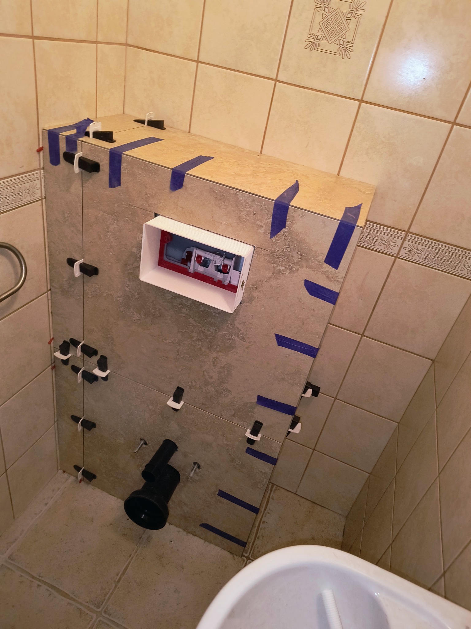 Płyty ceramiczne mocowane do obudowy stelaża podtynkowego toalety, z użyciem klipsów poziomujących i niebieskiej taśmy maskującej, widoczny fragment miski ustępowej.