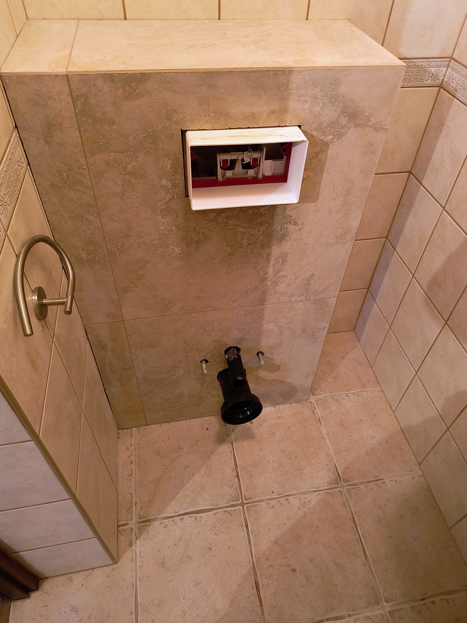 Ściana w beżowych płytkach z zamontowanym stelażem podtynkowym WC i wystającymi rurami kanalizacyjnymi, widok z dołu.