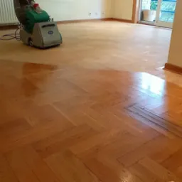 Cyklinowanie parkietu w jodełkę: maszyna do cyklinowania w trakcie pracy, widoczna r&oacute;żnica w kolorze między obrobioną a surową częścią podłogi.