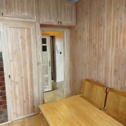 Wykończone drewnem wnętrze: boazeria ścienna, zabudowa szafkowa i stół z krzesłami. Widoczny parkiet ułożony w jodełkę i fragment ściany z brązowych płytek.