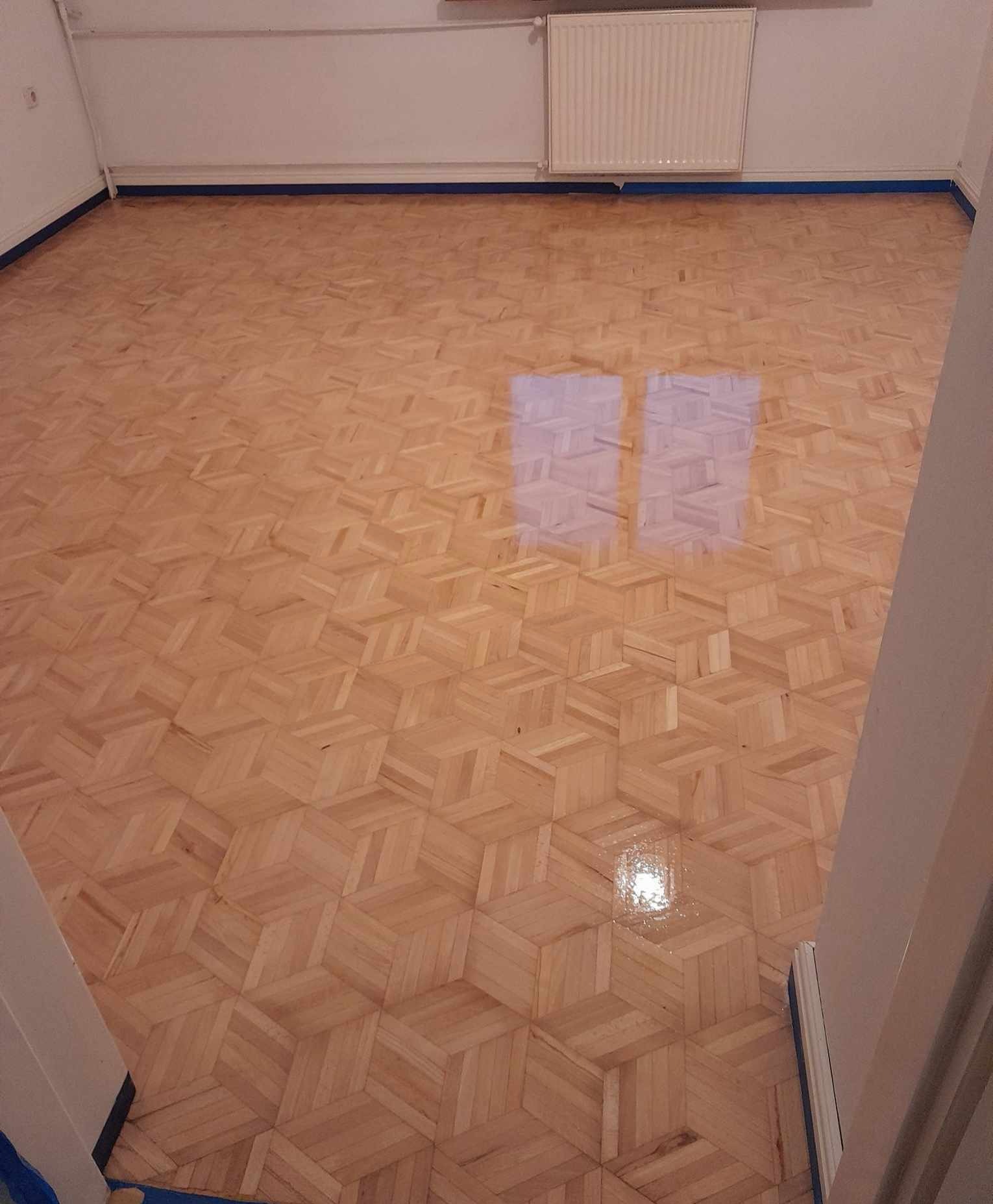 Błyszcząca, odnowiona podłoga parkietowa w jodełkę, zabezpieczona niebieską taśmą malarską przy ścianach. Widoczne odbicie okna w lakierowanej powierzchni.