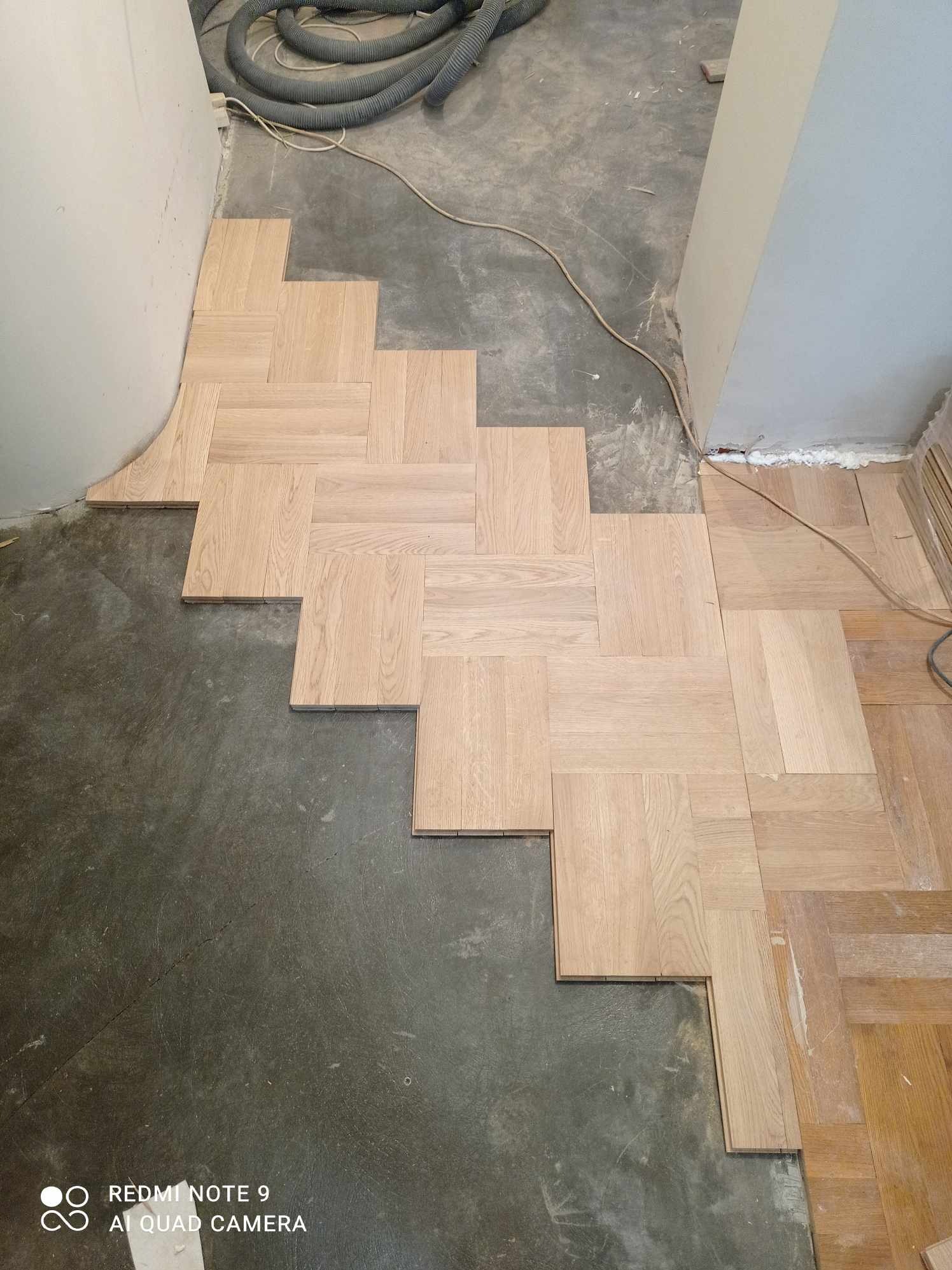 Układanie parkietu w jodełkę francuską na surowym podłożu. Częściowo ułożona podłoga z jasnego drewna. Widoczny odkurzacz przemysłowy i przewody.