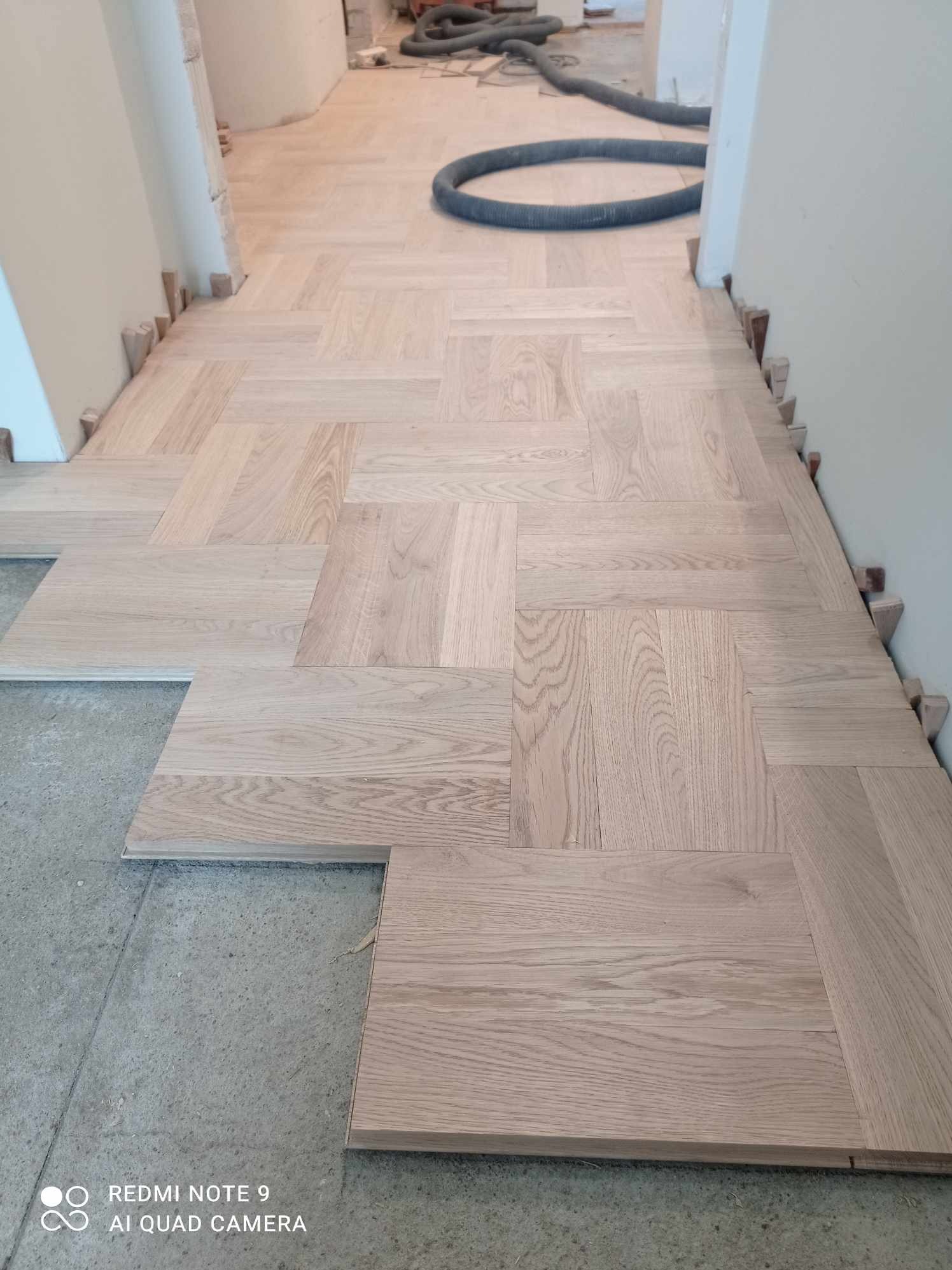 Parkiet ułożony we wzór jodełki, z przerwami na krawędziach i drewnianymi klinami dystansowymi przy ścianie. W tle wąż od odkurzacza przemysłowego.