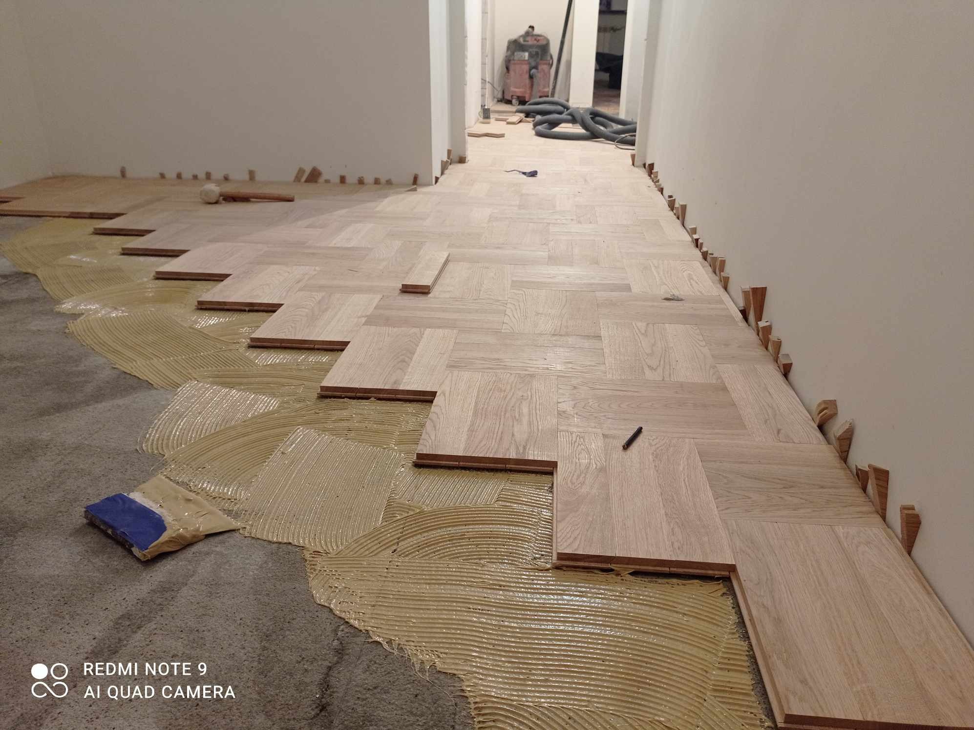 Układanie parkietu w jodełkę: klej na podłodze, fragment ułożonej podłogi, dylatacja przy ścianie z klinami. W głębi pomieszczenia maszyna do obróbki podłóg.