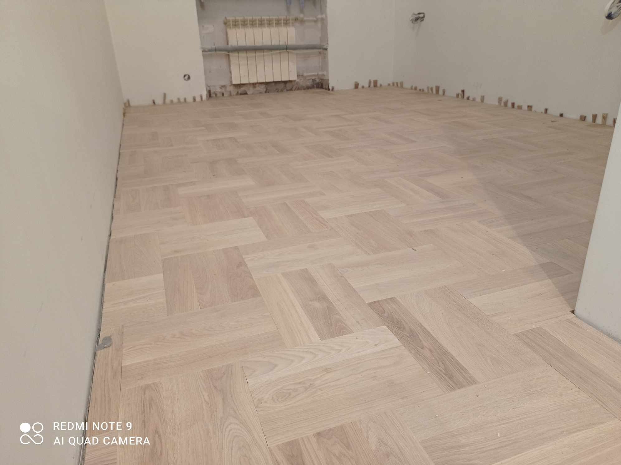 Jasny, ułożony w jodełkę parkiet w pokoju z grzejnikiem i listwami przypodłogowymi. Widoczne kliny dystansowe przy ścianach.