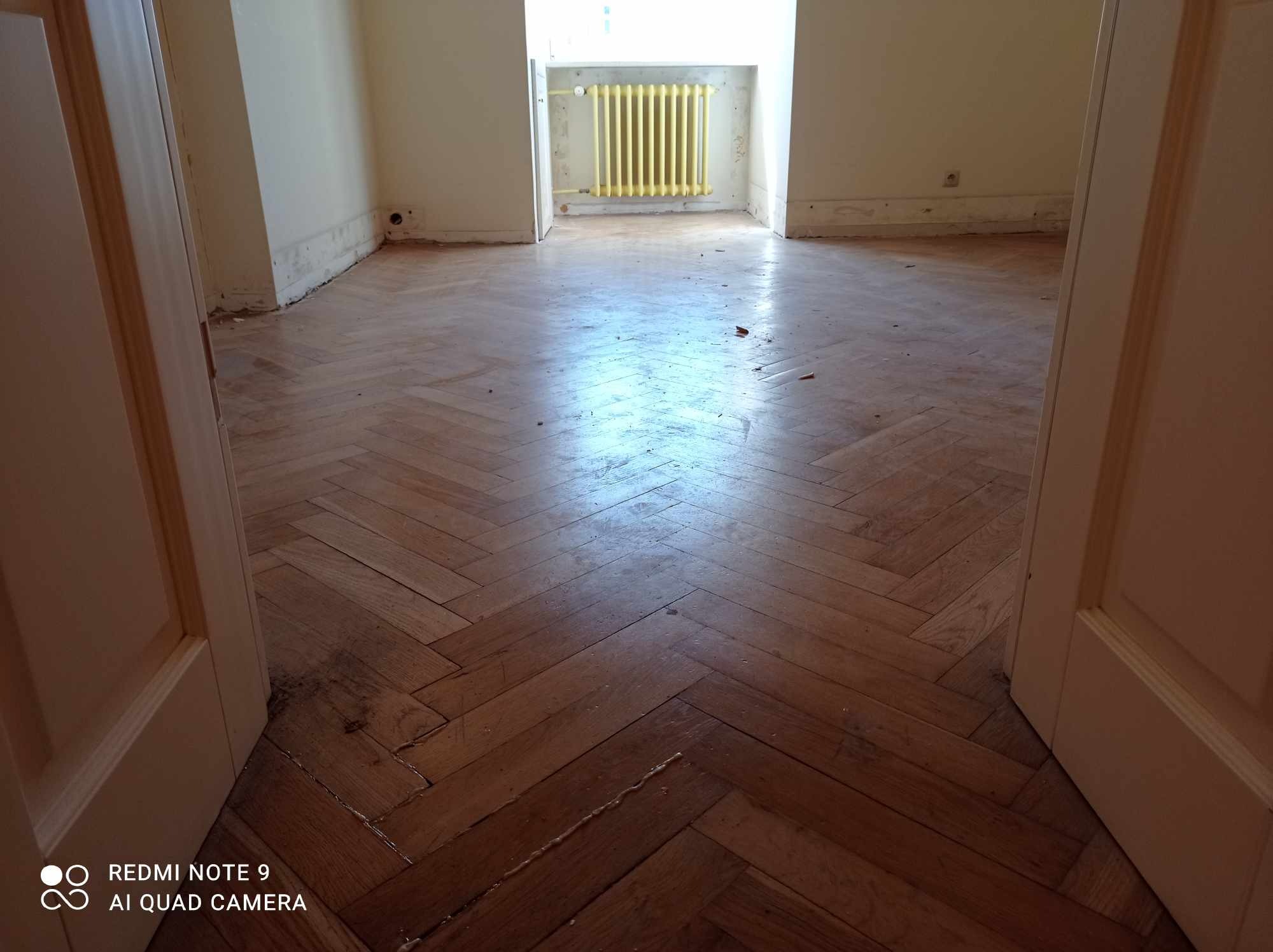 Parkiet ułożony w jodełkę, widoczny z perspektywy wejścia do pokoju. Żółty grzejnik w tle. Podłoga wymaga renowacji, widoczne ślady użytkowania i zabrudzenia.
