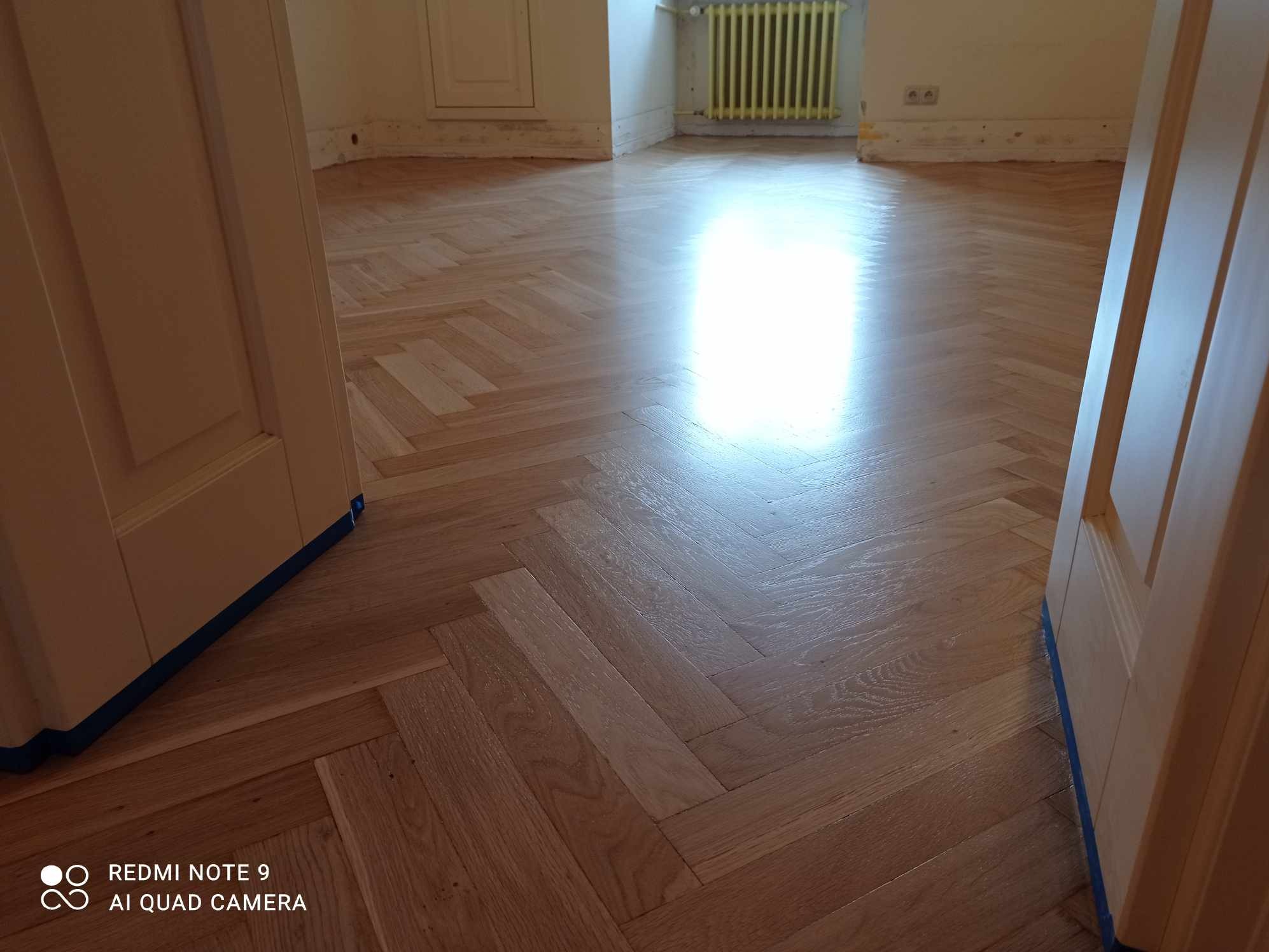 Błyszcząca, jasna podłoga z parkietu ułożonego we wzór jodełki, widoczna przez otwarte drzwi. Świeżo położona podłoga odbija światło. Zabezpieczone listwy przypodłogowe.