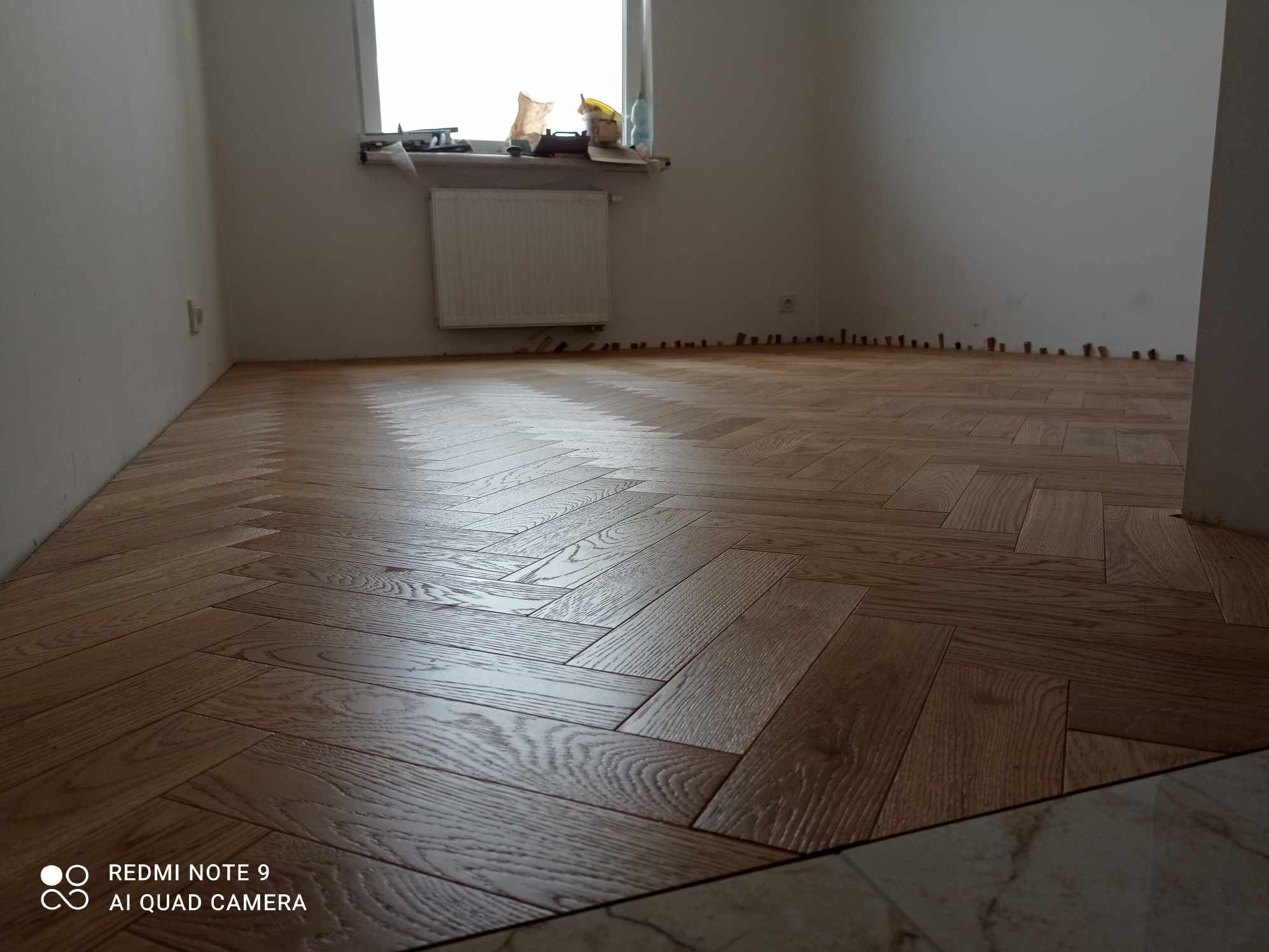 Parkiet ułożony w jodełkę, widoczne listwy przypodłogowe do zamontowania. Na parapecie okna leżą narzędzia.