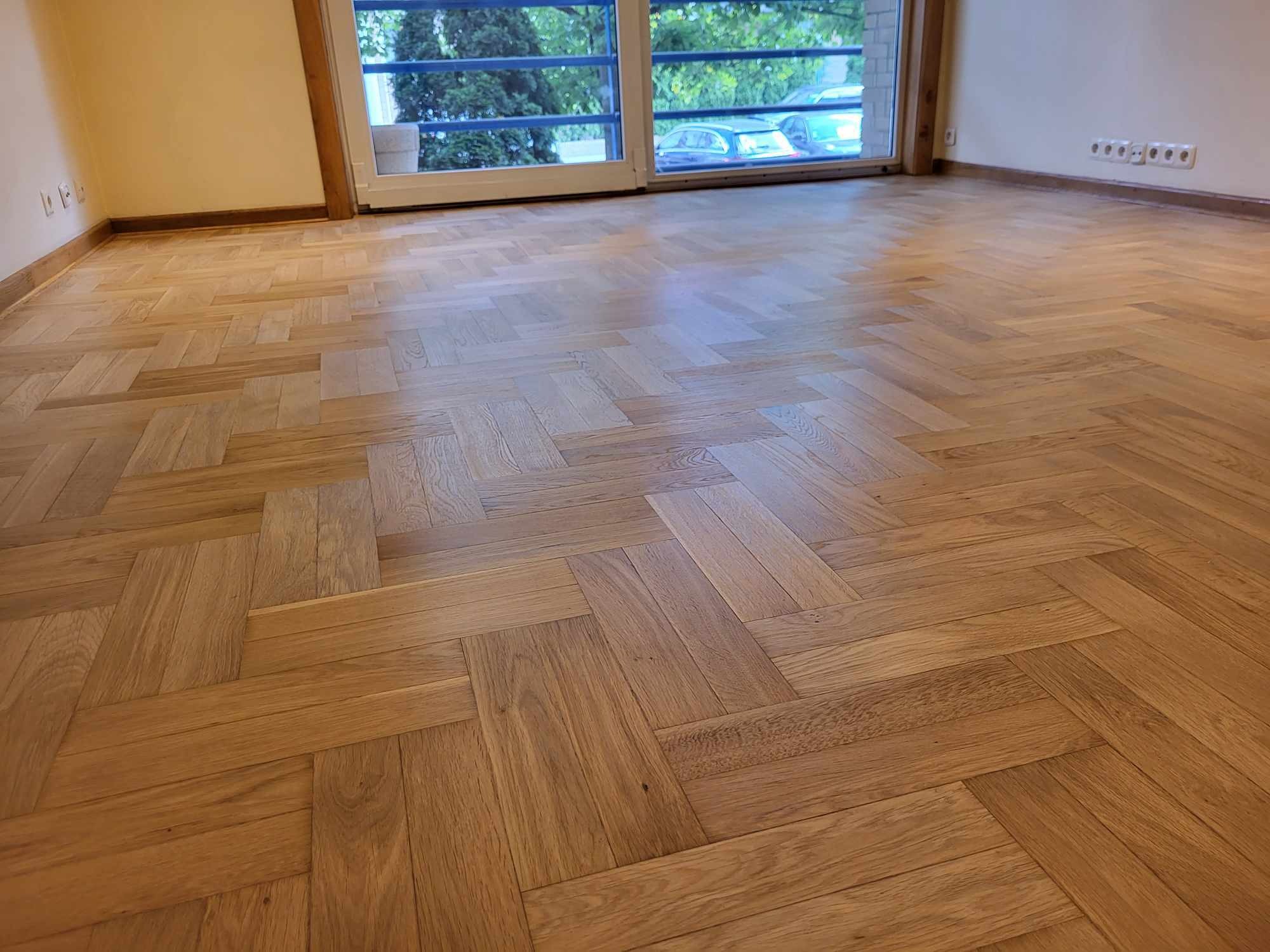 Jasny parkiet ułożony we wzór jodełki w przestronnym pokoju z oknem balkonowym i listwami przypodłogowymi. Widoczne gniazdka elektryczne na ścianie.