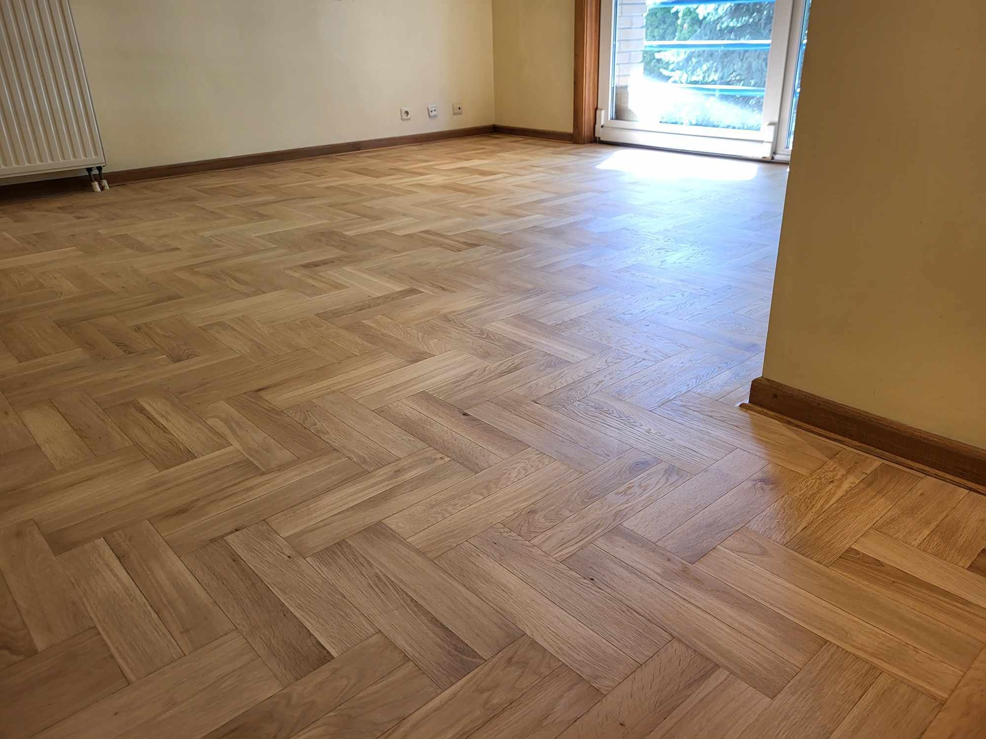 Jasny parkiet ułożony w jodełkę w przestronnym pokoju z kremowymi ścianami i białymi drzwiami balkonowymi. Widoczne listwy przypodłogowe.