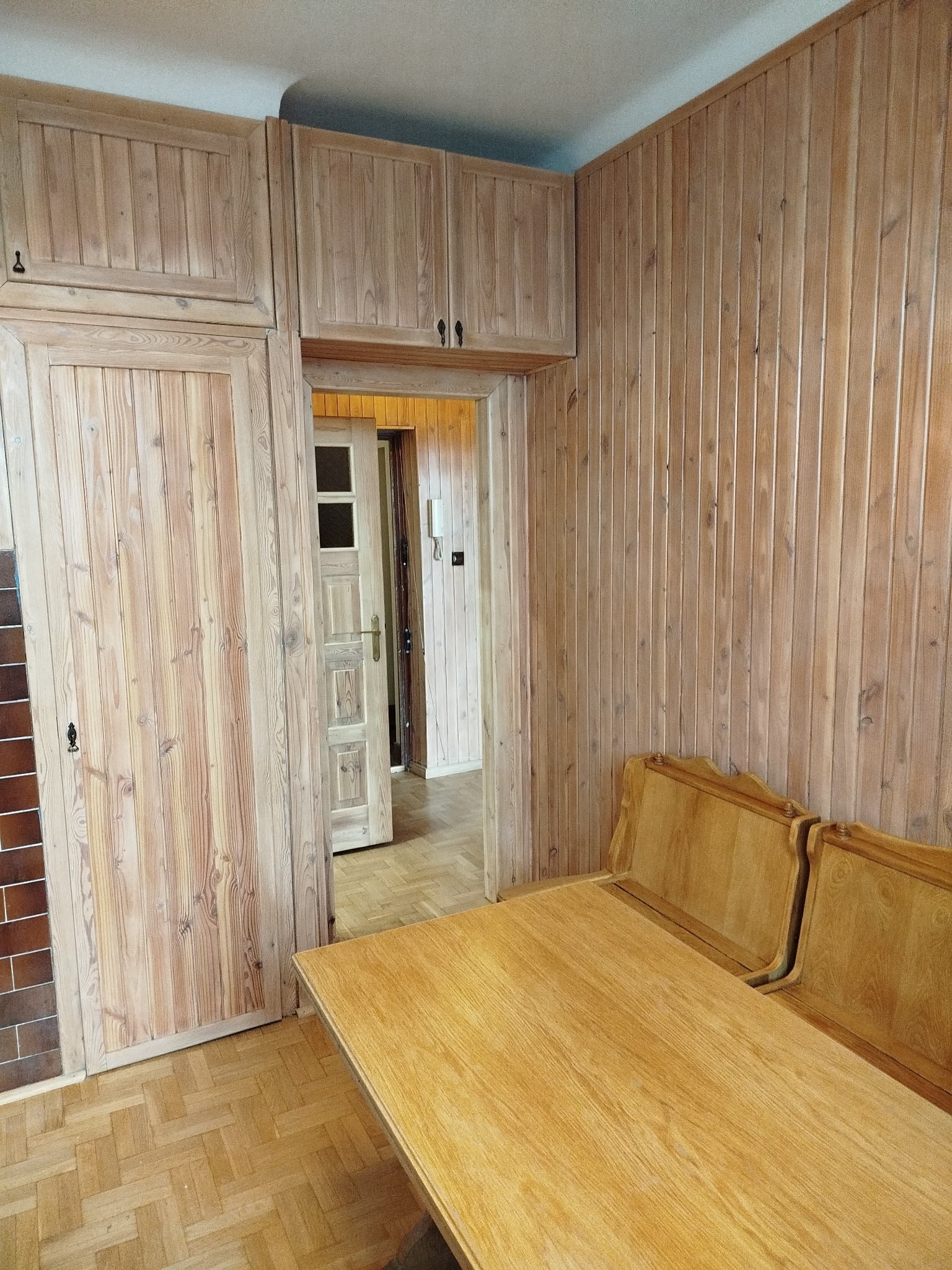 Wykończone drewnem wnętrze: boazeria ścienna, zabudowa szafkowa i stół z krzesłami. Widoczny parkiet ułożony w jodełkę i fragment ściany z brązowych płytek.