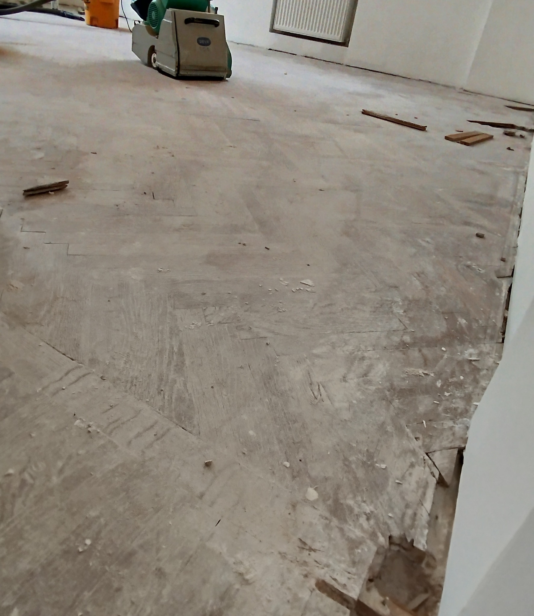 Parkiet w trakcie renowacji, widoczna maszyna do cyklinowania i resztki starej powłoki, przygotowanie podłoża do dalszych prac wykończeniowych.