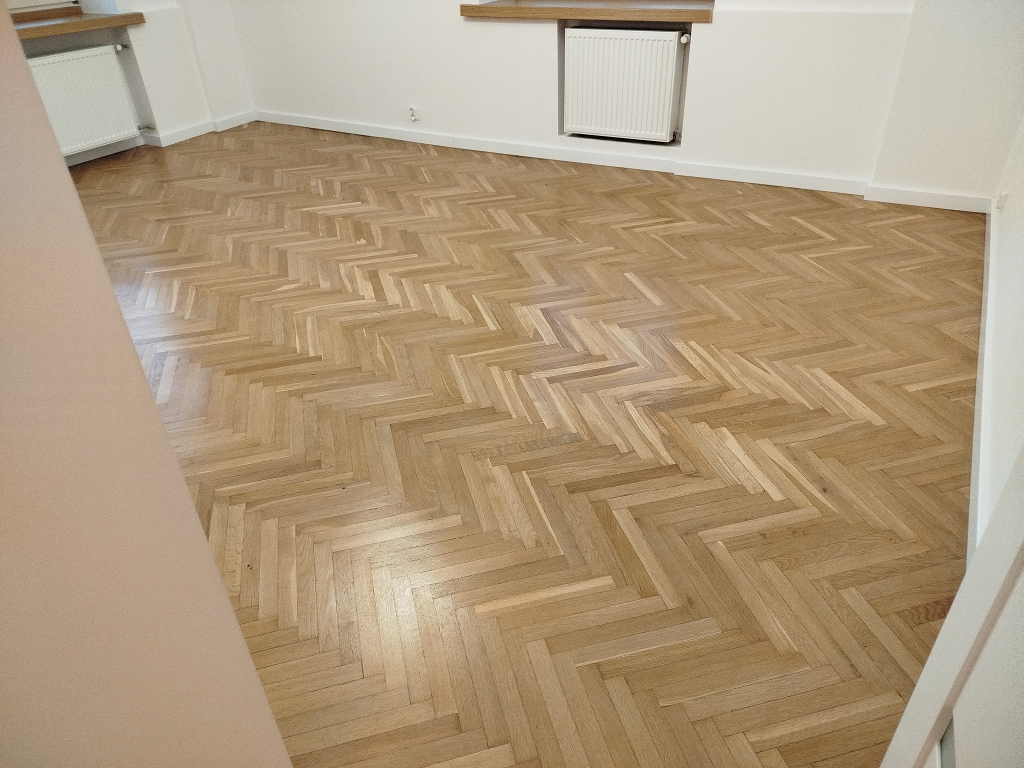 Jasny parkiet ułożony w jodełkę w świeżo wyremontowanym pokoju z białymi ścianami i grzejnikiem. Widok na całą podłogę, brak mebli.