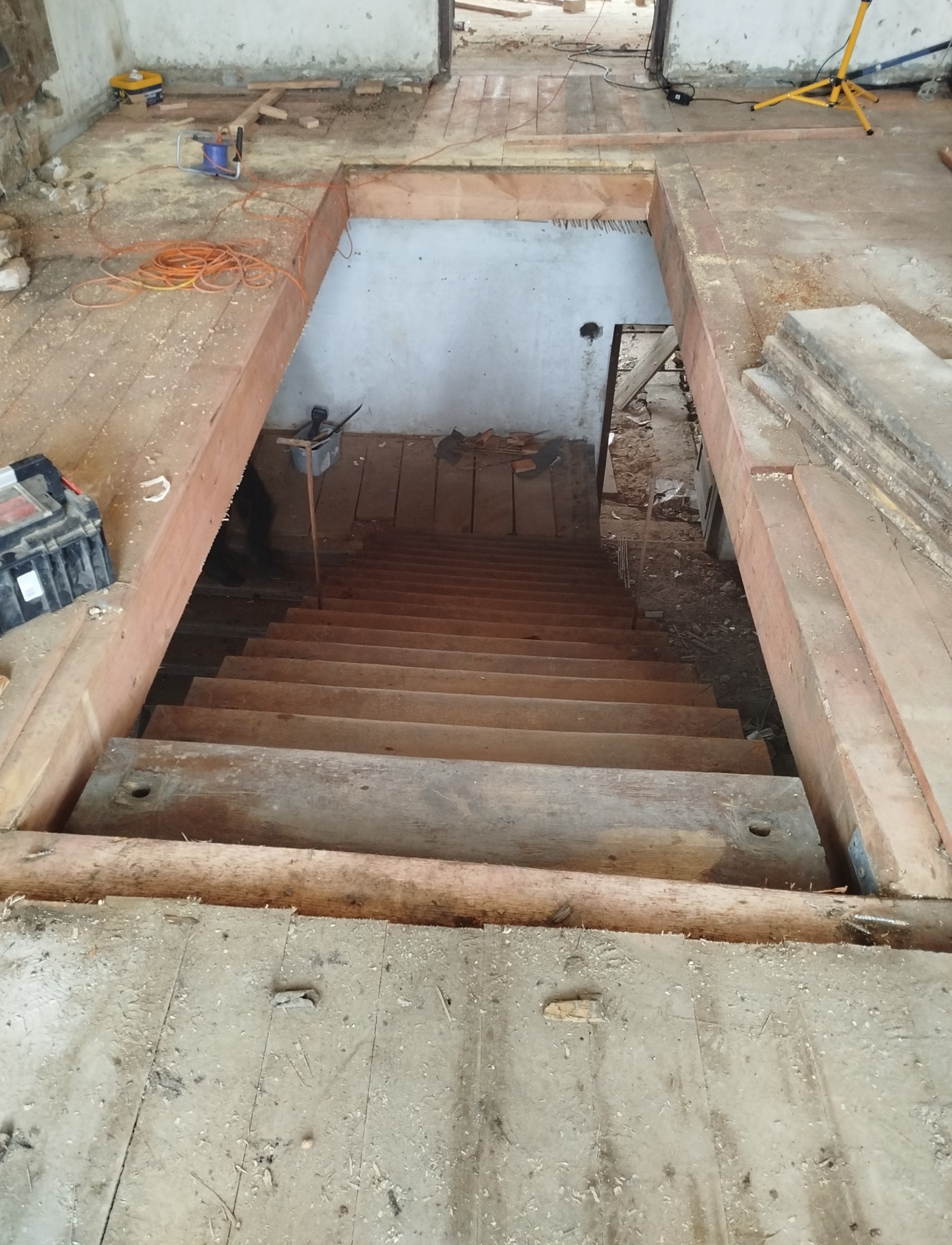 Demontaż drewnianych schodów w trakcie remontu. Widoczne narzędzia, kable i deski. Podłoga pokryta pyłem i resztkami materiałów. Ujęcie z góry.