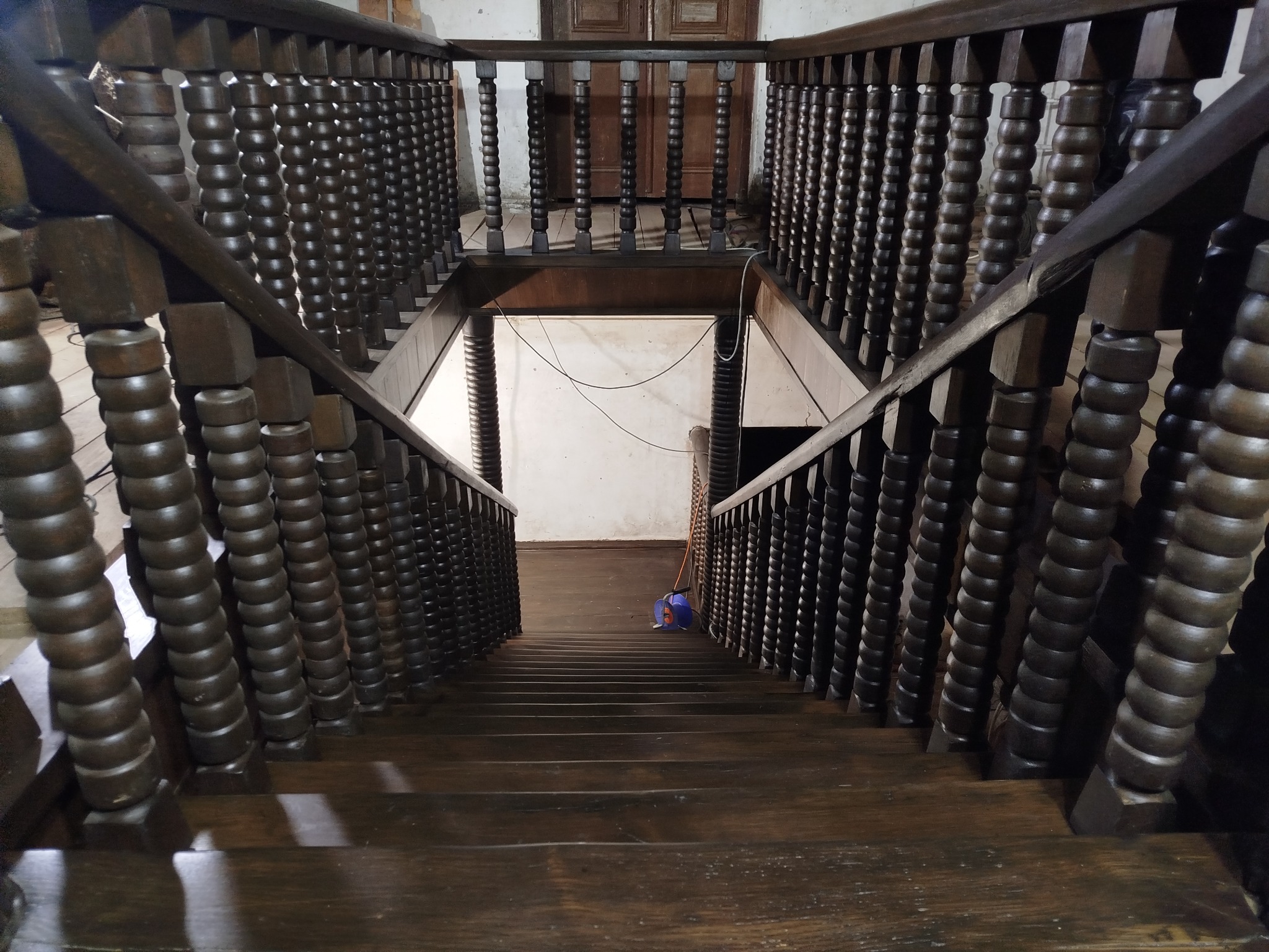 Ciemne, drewniane schody z toczonymi balustradami, widziane z góry. Na podeście widoczny sprzęt. Ujęcie podkreśla perspektywę i detale stolarstwa.