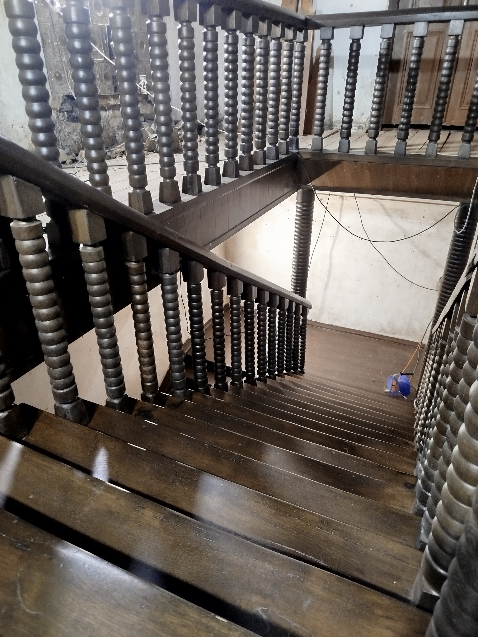 Drewniane schody z toczonymi balustradami, widok z góry. Na podłodze widoczny sprzęt do sprzątania. Charakterystyczne zdobienia balustrad.