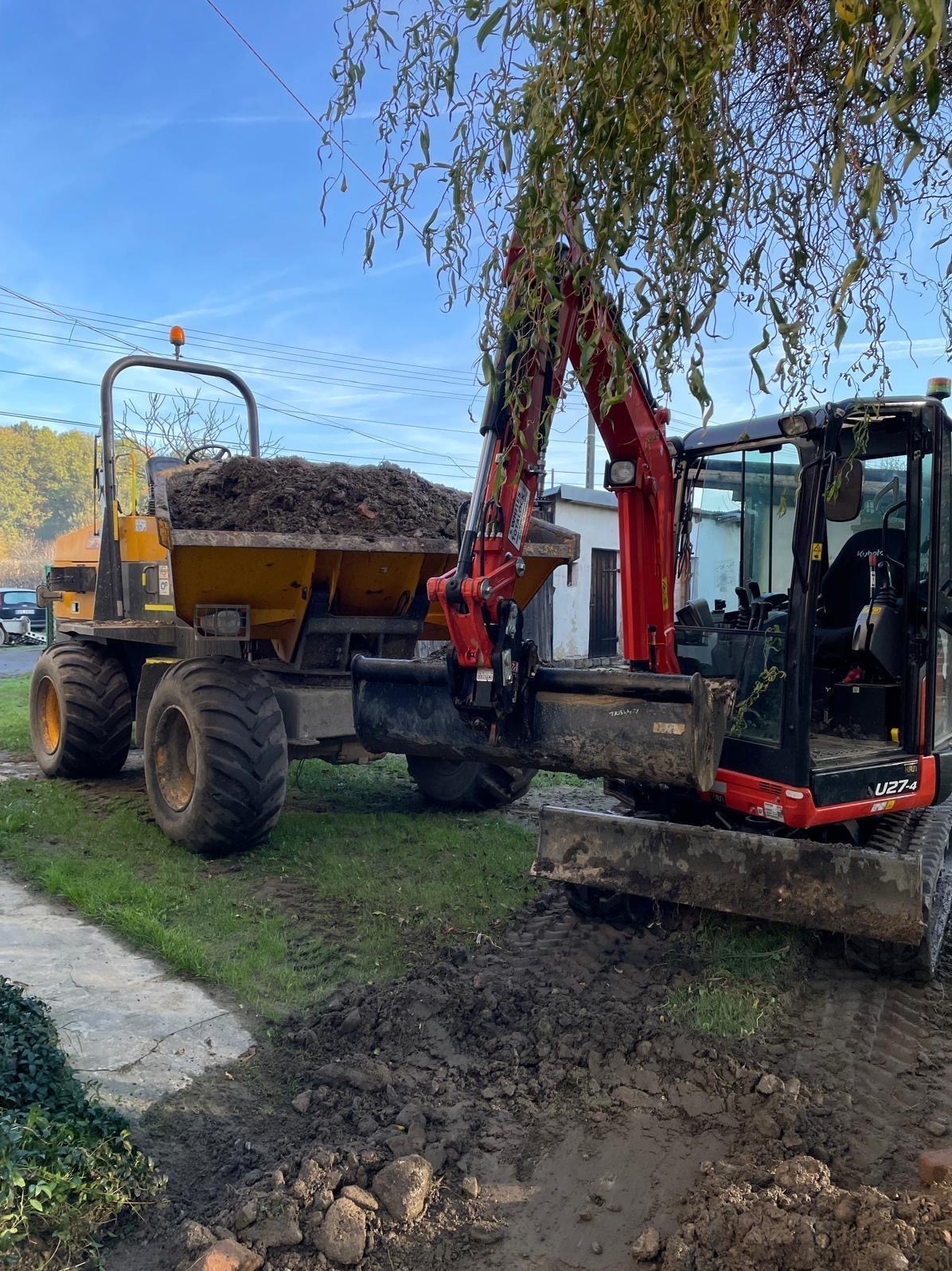 Żółta wywrotka wypełniona ziemią i czerwona mini koparka Kubota U27-4 podczas prac ziemnych na trawiastym terenie w pobliżu chodnika i drzewa.
