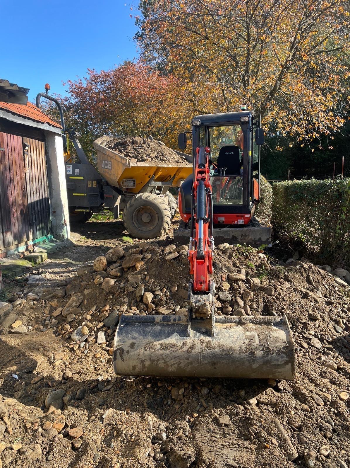 Czerwony mini-koparka Kubota z łyżką na pierwszym planie, w tle wywrotka JCB wypełniona ziemią, jesienne drzewa i drewniany budynek.