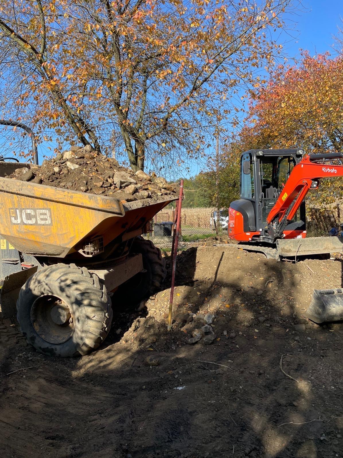 Żółta wywrotka JCB wypełniona ziemią i kamieniami obok czerwonej mini koparki Kubota na placu budowy z drzewami w tle, jesienne liście, słoneczny dzień.