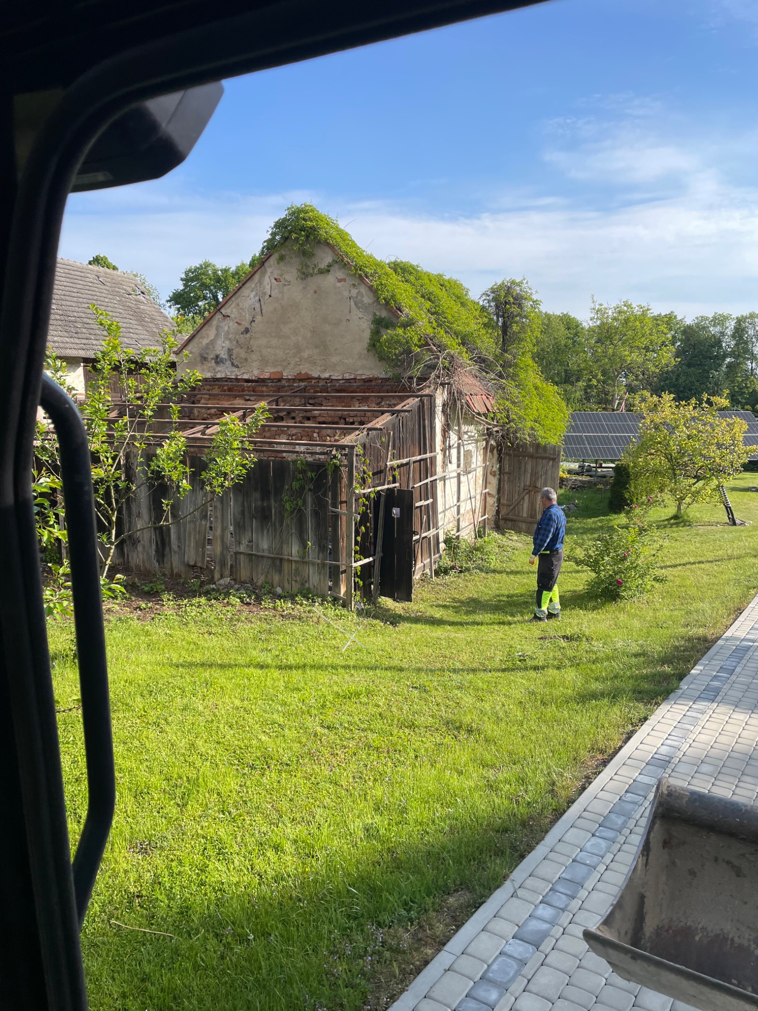 Mężczyzna w odblaskowej kamizelce stoi obok starego, częściowo rozebranego budynku z ceglanym szczytem porośniętym zielenią. W tle panele słoneczne.
