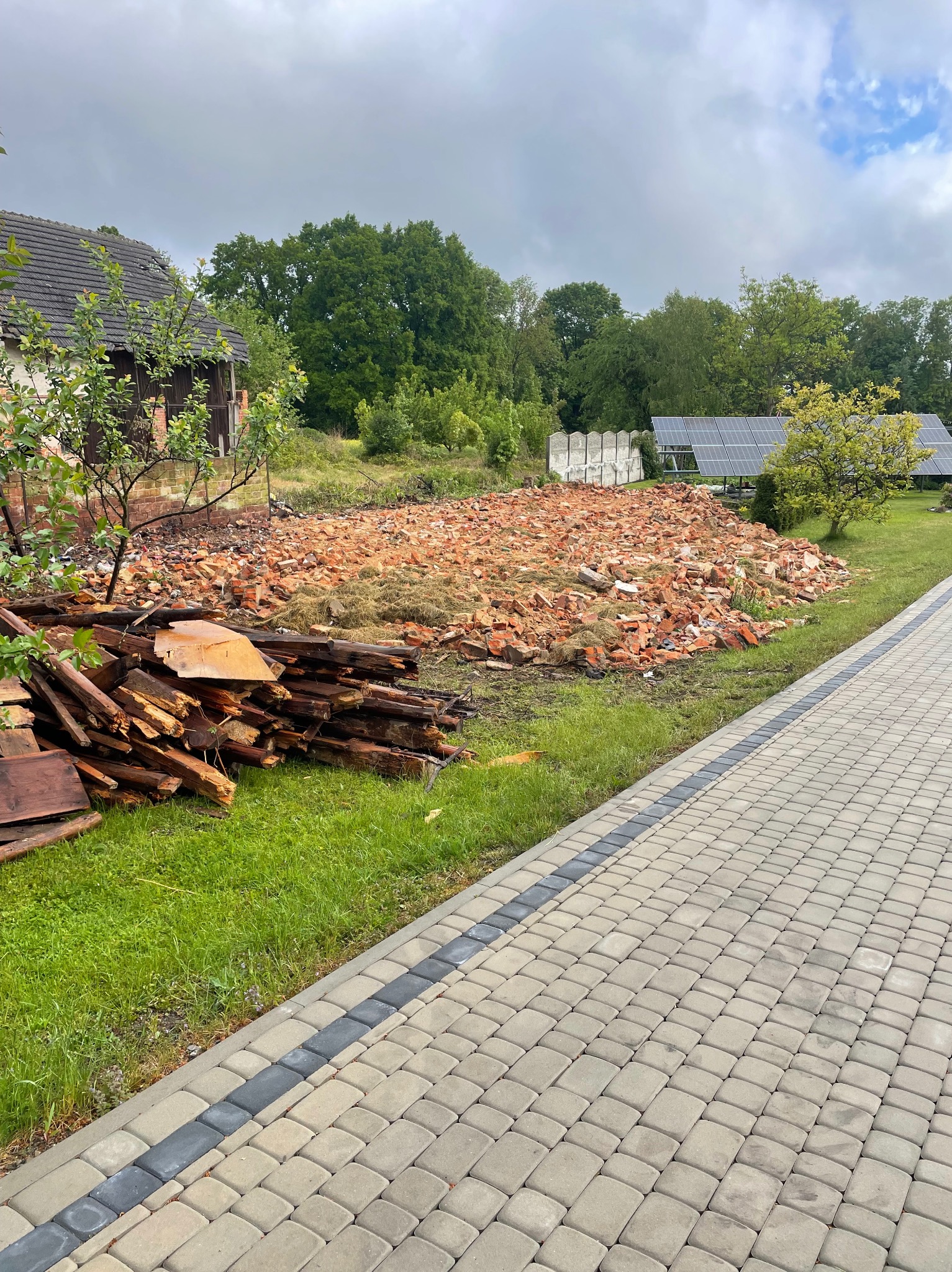 Teren po rozbiórce budynku z gruzem ceglanym i stertą drewna obok chodnika z kostki, w tle panele słoneczne i zieleń. Pochmurne niebo.