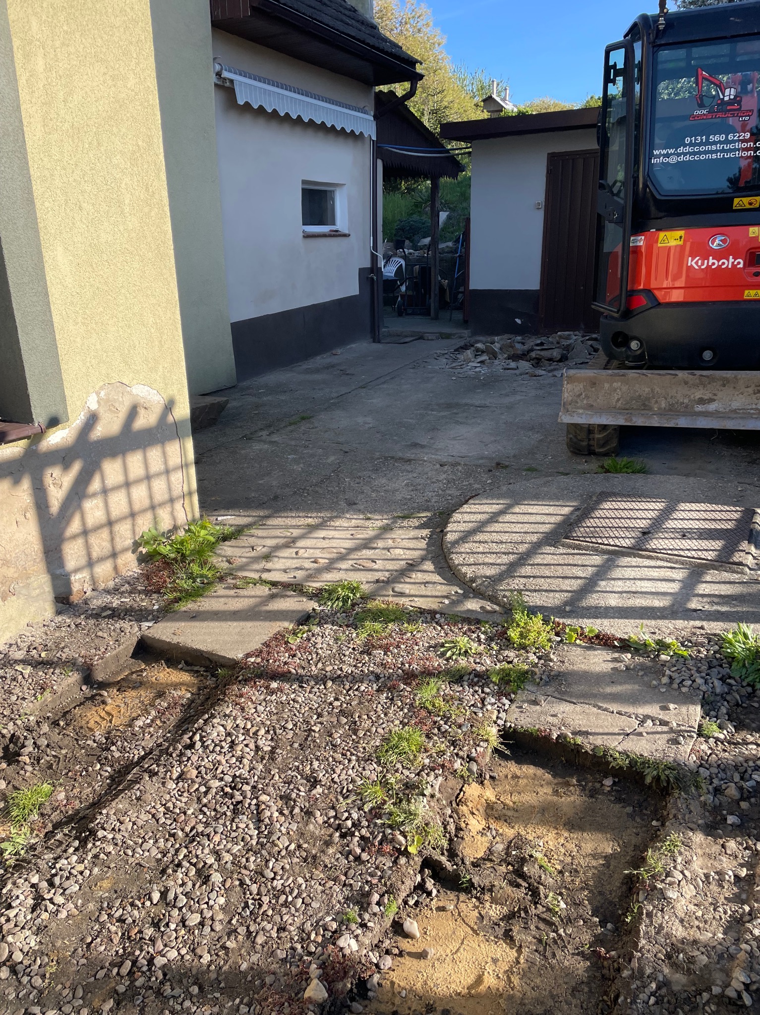Wykop pod fundamenty z widocznym fragmentem mini koparki Kubota. Fragmenty betonowych płyt chodnikowych i gruzu. Cień kraty na elewacji budynku.