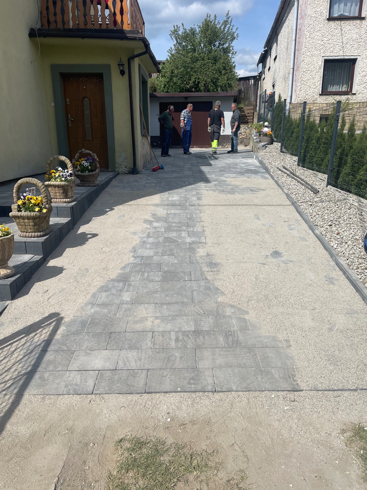 Ułożona szara kostka brukowa na podjeździe, z widocznymi śladami piasku i czterema osobami w tle, stojącymi obok budynku z jasną elewacją.
