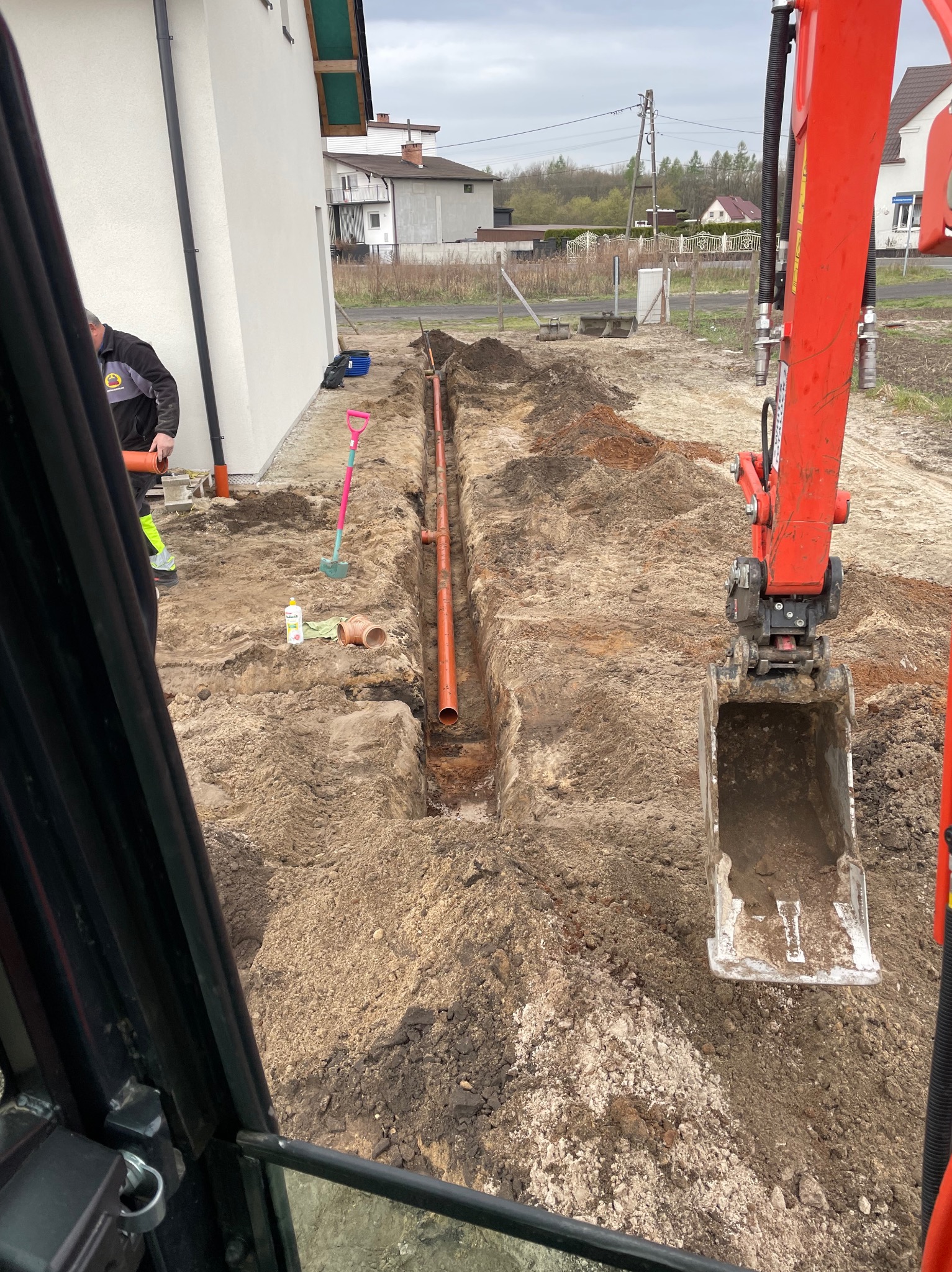Pomarańczowe rury ułożone w wykopie ziemnym obok budynku, widok z kabiny koparki. Pracownik łączy rury, obok leżą narzędzia.