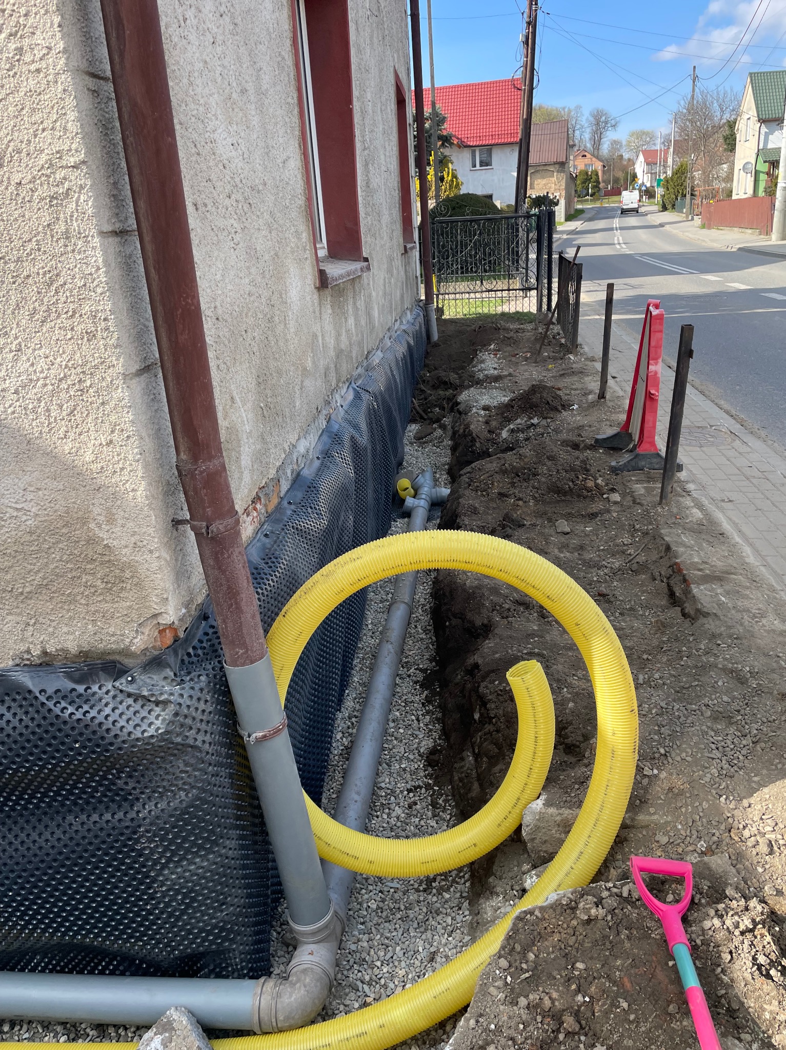 Odkopane fundamenty domu z widoczną izolacją i żółtą rurą drenażową. W tle ulica z domami i błękitne niebo. Mała, różowa łopatka leży na ziemi obok wykopu.