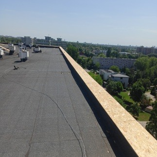 Płaski dach pokryty papą z drewnianą obróbką krawędziową na tle zieleni miejskiej i błękitnego nieba. Widok z góry, perspektywa zbieżna, słoneczny dzień.