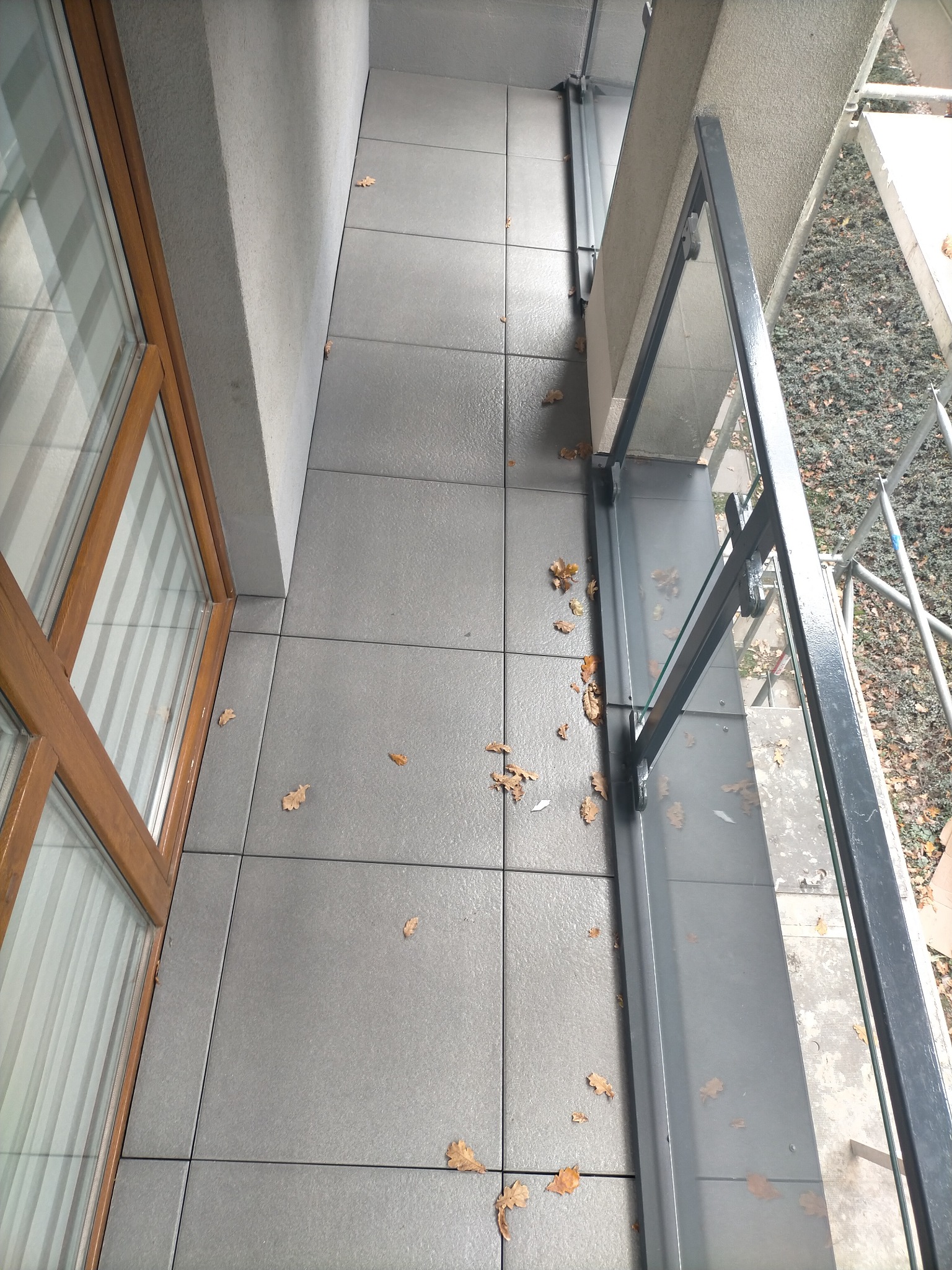 Balkon z szarą, nowoczesną terakotą i szklaną balustradą. Widok z góry, z widocznymi jesiennymi liśćmi. Ujęcie z wysokości, prezentuje wykończenie balkonu.