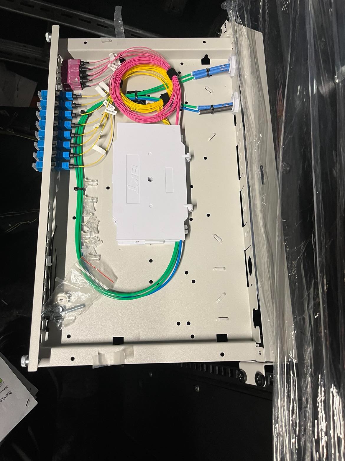 Rozdzielnica światłowodowa BKT z kolorowymi kablami światłowodowymi i złączami, częściowo zamontowana w Jarosławiu, widoczne śruby i elementy montażowe w woreczku.