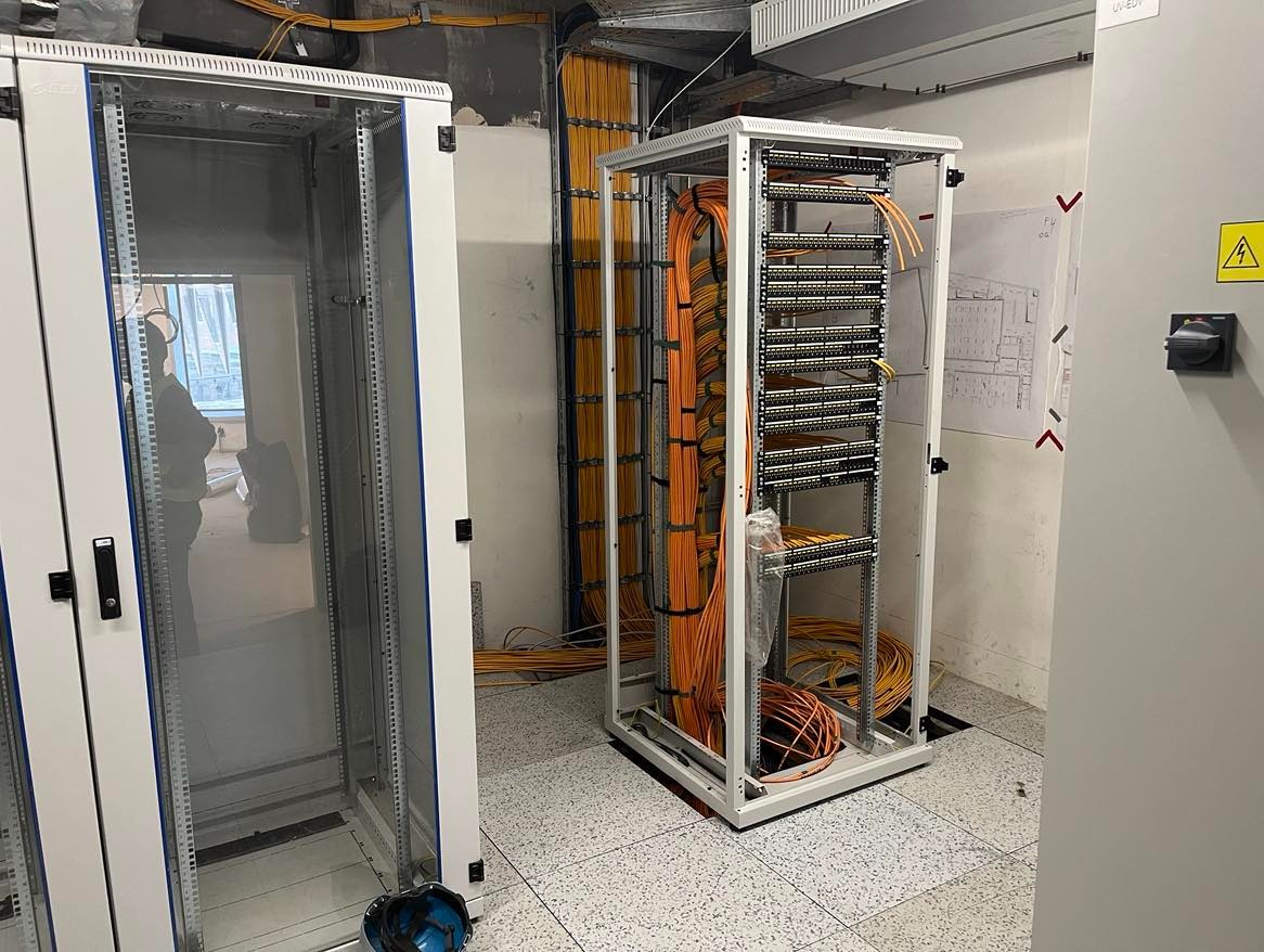 Dwie szafy serwerowe, jedna pusta z widocznym odbiciem osoby, druga z uporządkowanymi pomarańczowymi kablami, widoczne zakończenia kabli w listwach, na ścianie schemat instalacji.