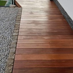 BLUE OAK SPÓŁKA Z OGRANICZONĄ ODPOWIEDZIALNOŚCIĄ - Widok z góry na drewniany taras z egzotycznego drewna Bangkirai, z kamiennym obrzeżem i szklaną balustradą, prowadzący w stronę domu.