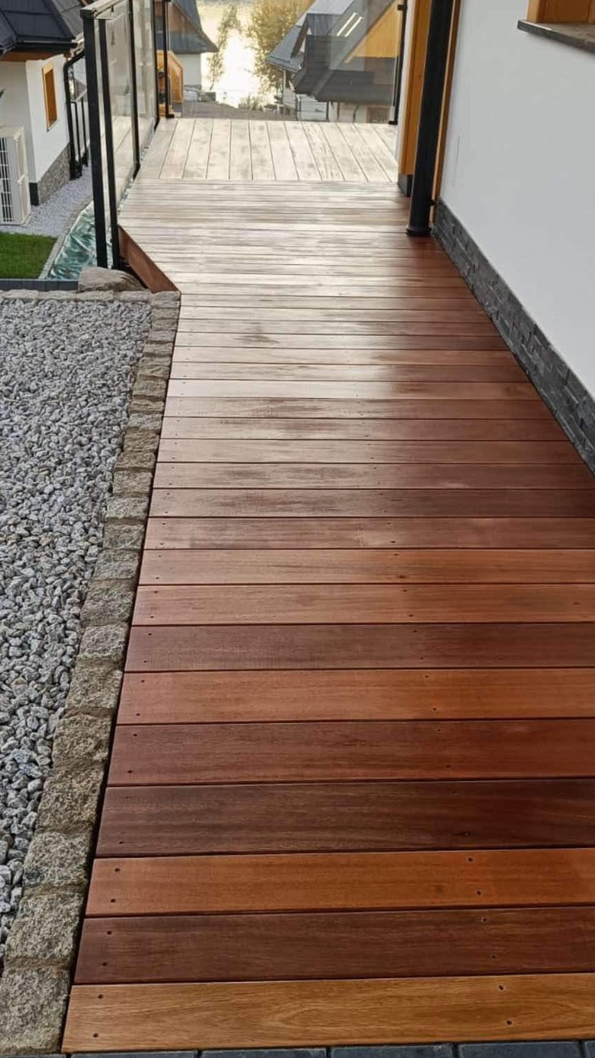 Widok z góry na drewniany taras z egzotycznego drewna Bangkirai, z kamiennym obrzeżem i szklaną balustradą, prowadzący w stronę domu.