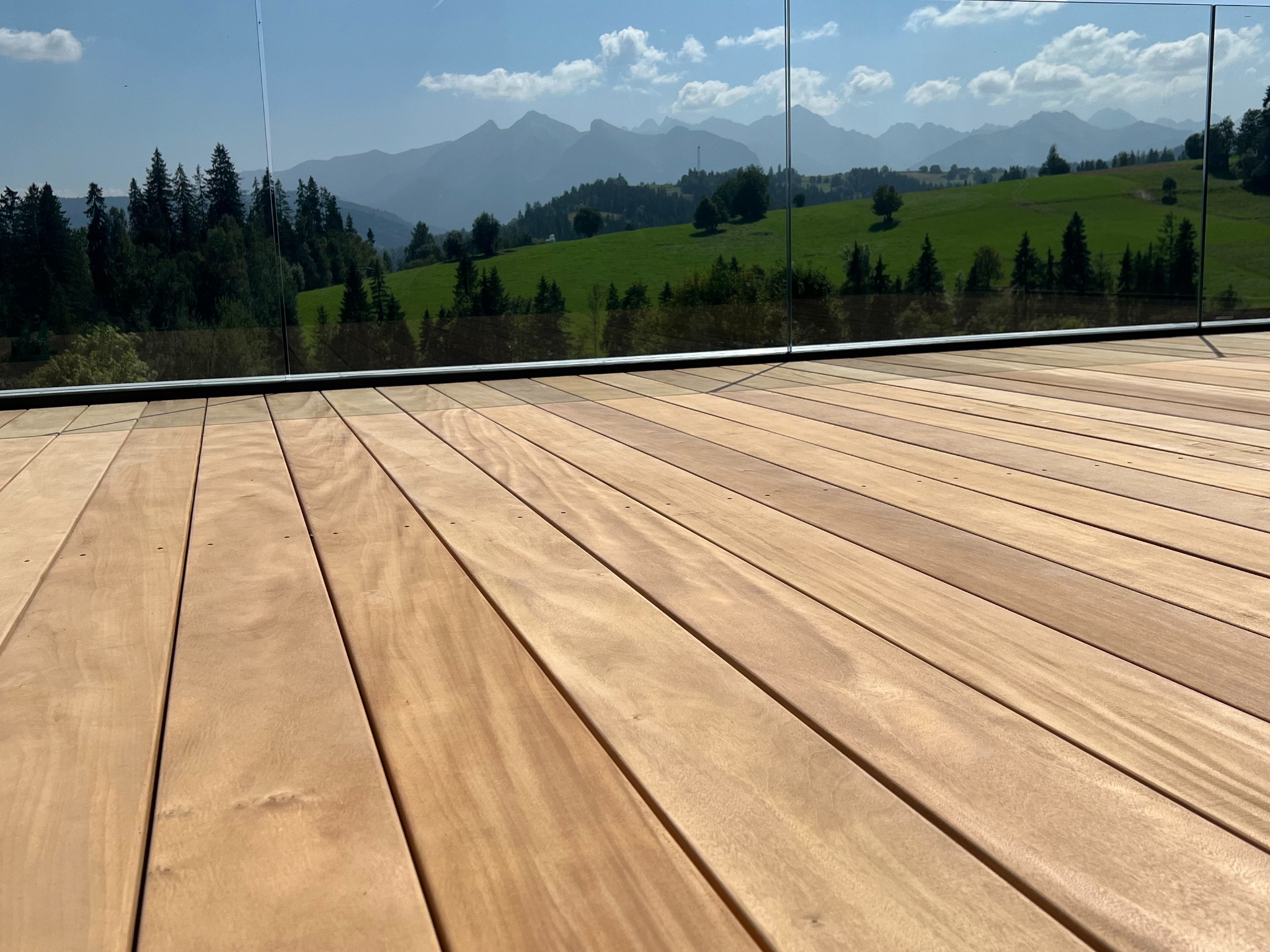 Gładka deska egzotyczna Garapa na tarasie z przeszkloną balustradą, widok na zielone wzgórza i szczyty górskie w tle.