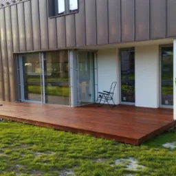 Nowoczesny dom z drewnianym tarasem, na trawniku stoi hulajnoga, na ścianie logo firmy 'Drewno dla domu'.